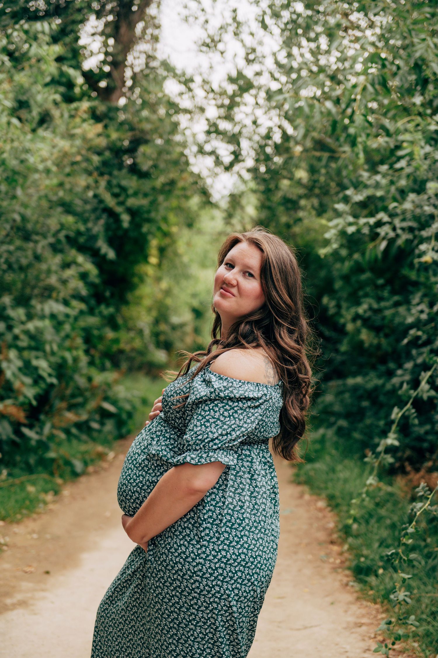 Pregnant woman in teal floral off-shoulder dress standing on a forest path, cradling her baby bump.