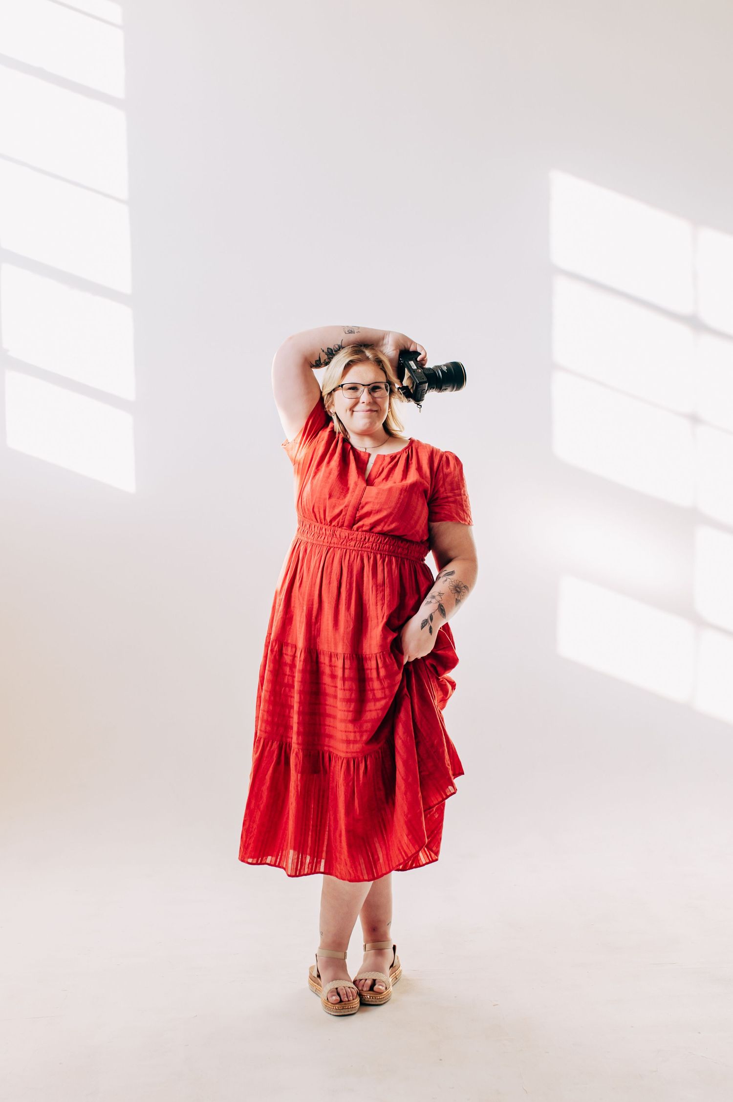 Woman in red dress holding camera aloft in bright white studio, wearing glasses and wedge sandals.