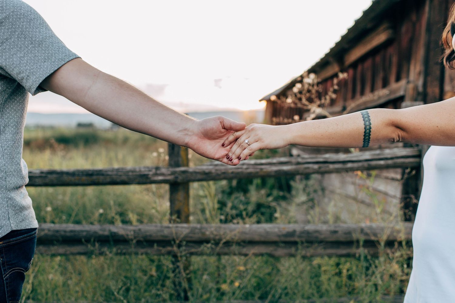 A couple holds hands tenderly near a rustic wooden fence surrounded by lush green grass in a romantic outdoor setting.