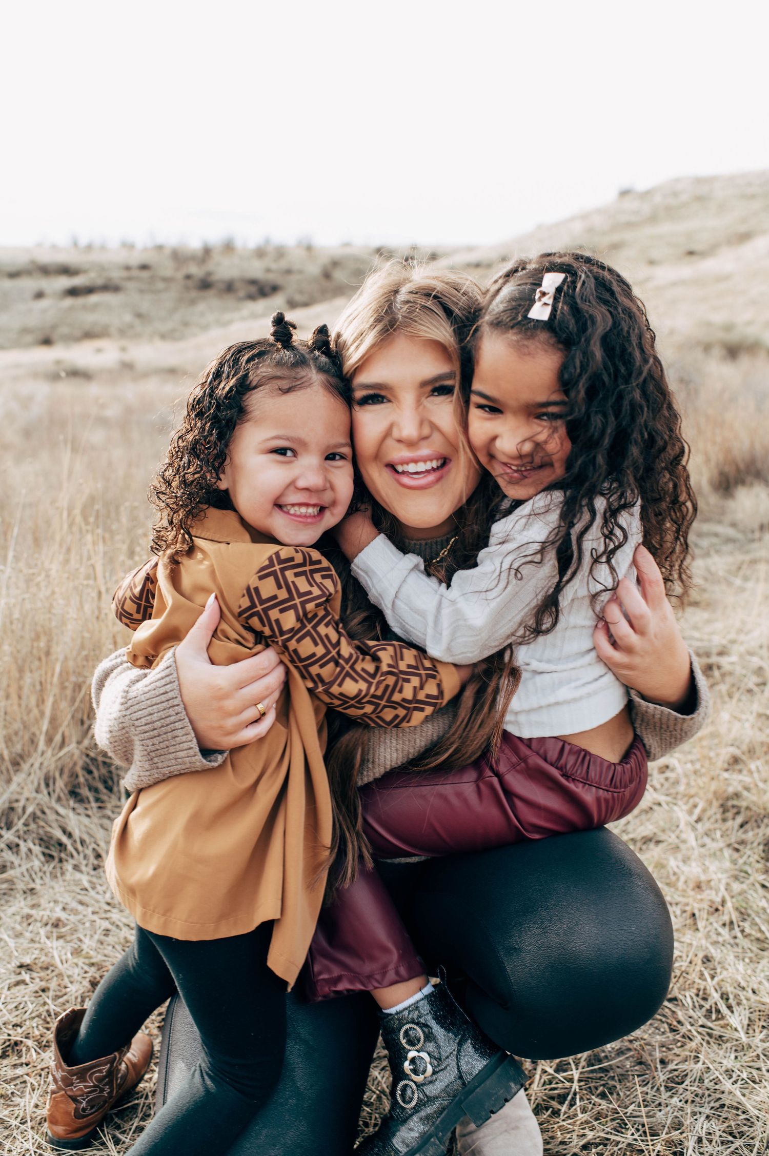 A smiling woman crouches outdoors hugging two curly-haired girls in a dry grassy field during fall.