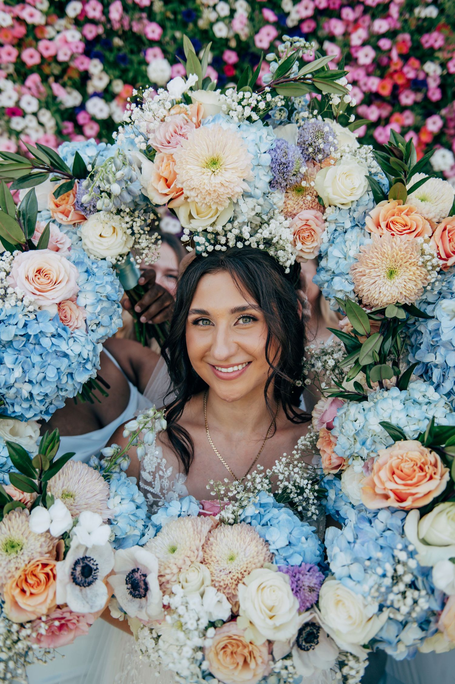 Smiling bride surrounded by lush pastel floral arrangement featuring blue hydrangeas, peach roses, and white blooms.
