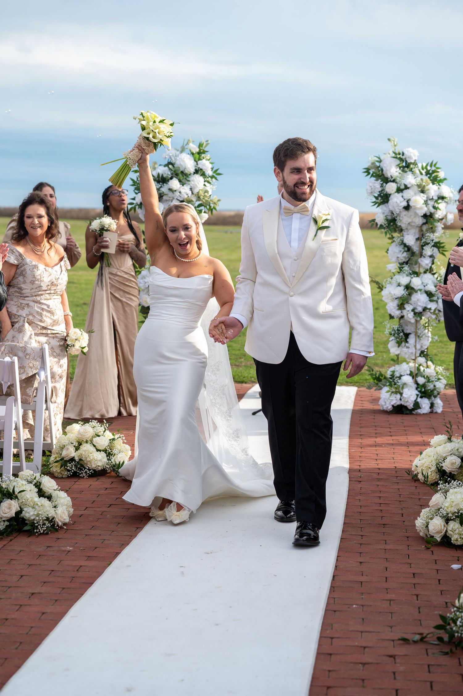 A wedding couple and guests walk down a red brick aisle decorated with white floral arrangements at an outdoor ceremony.