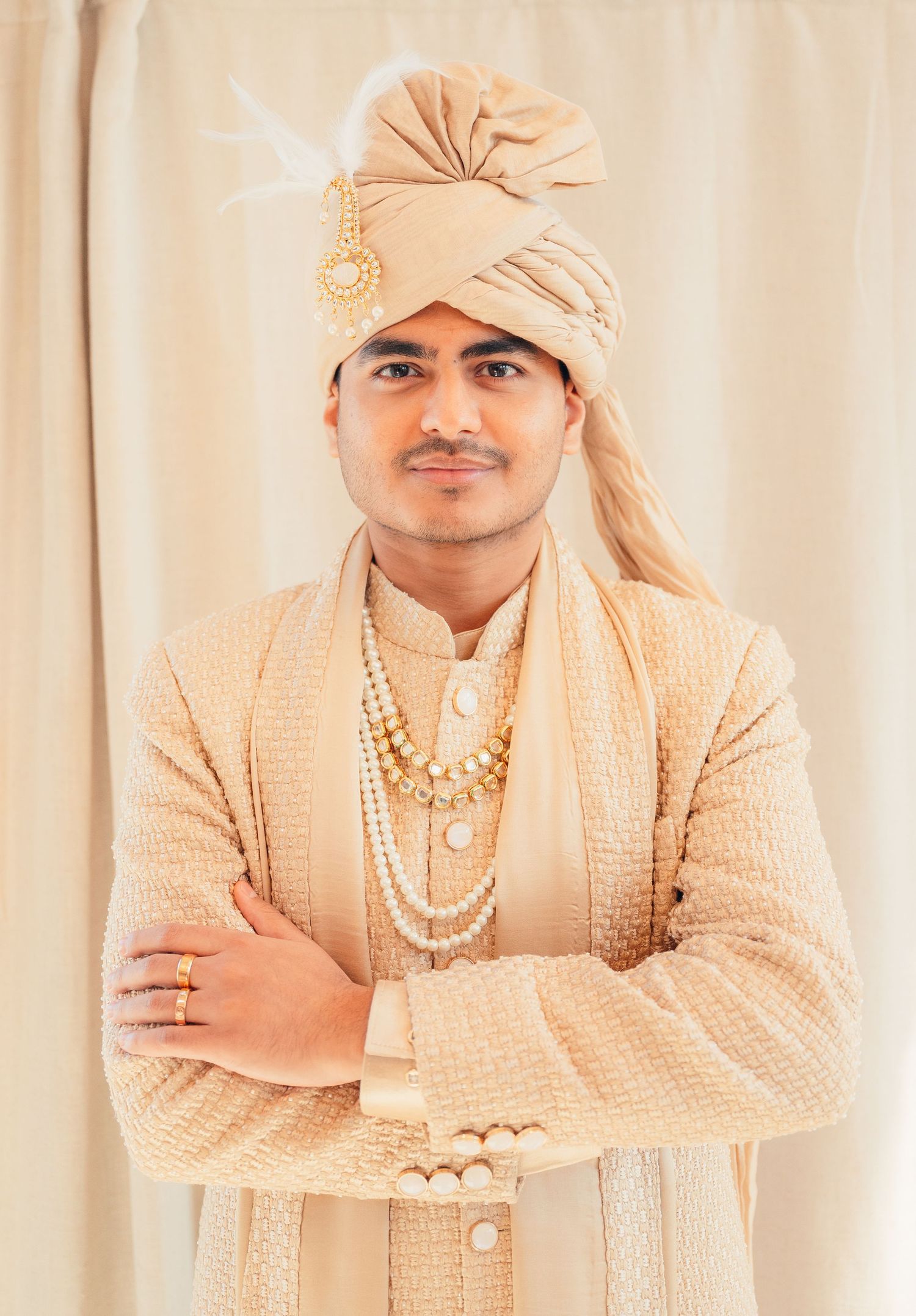 Traditional groom's portrait in cream-colored sherwani and pearl necklaces against light backdrop.
