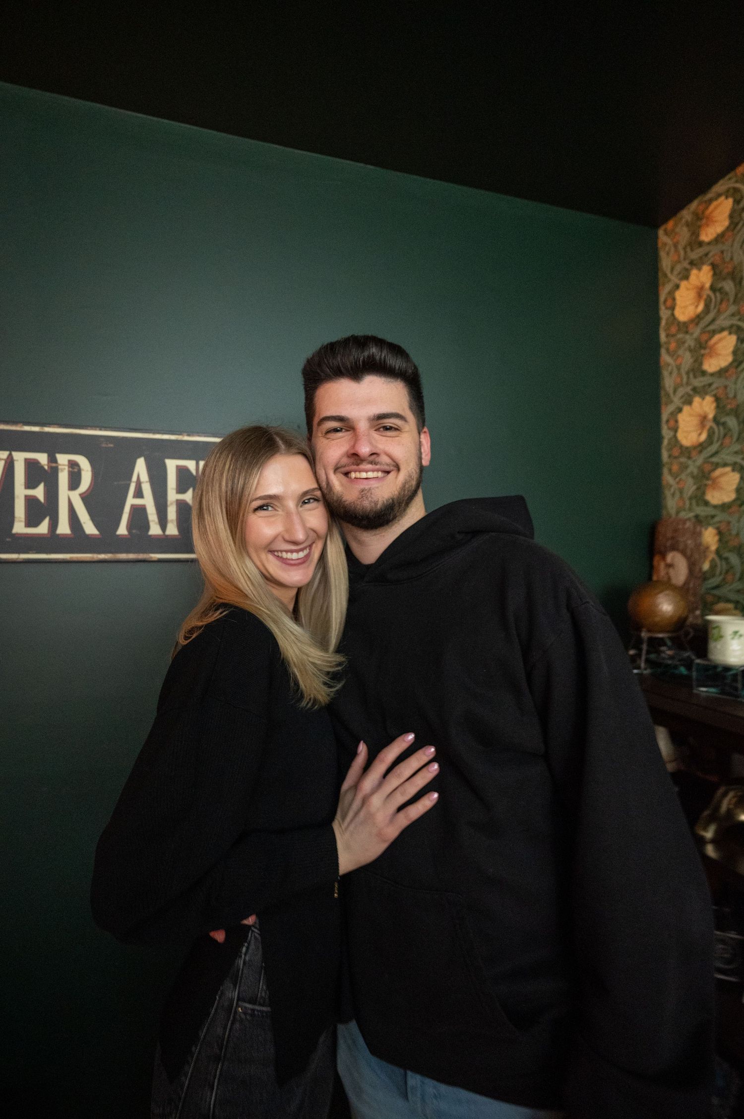 A couple embraces and smiles in front of a green wall with decorative text.