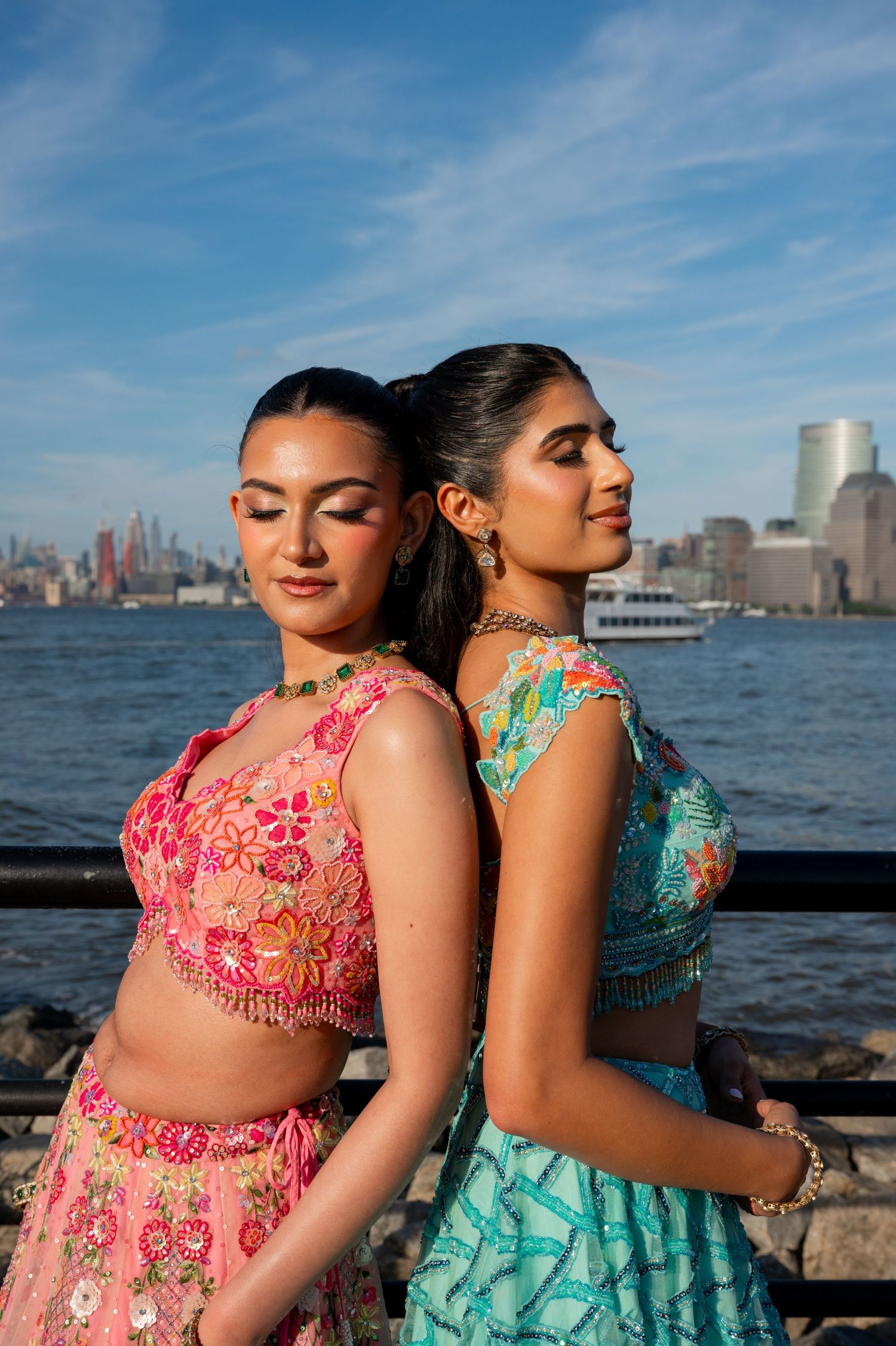 Two women in colorful formal dresses pose against NYC skyline at waterfront during sunset.
