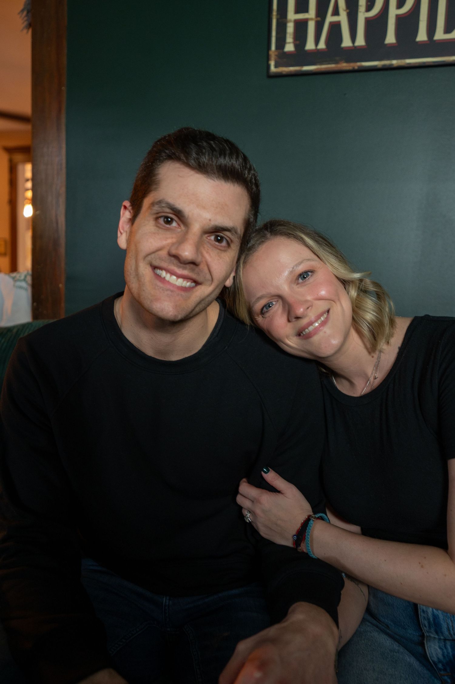 A couple in matching black outfits smiles warmly in front of a green wall.