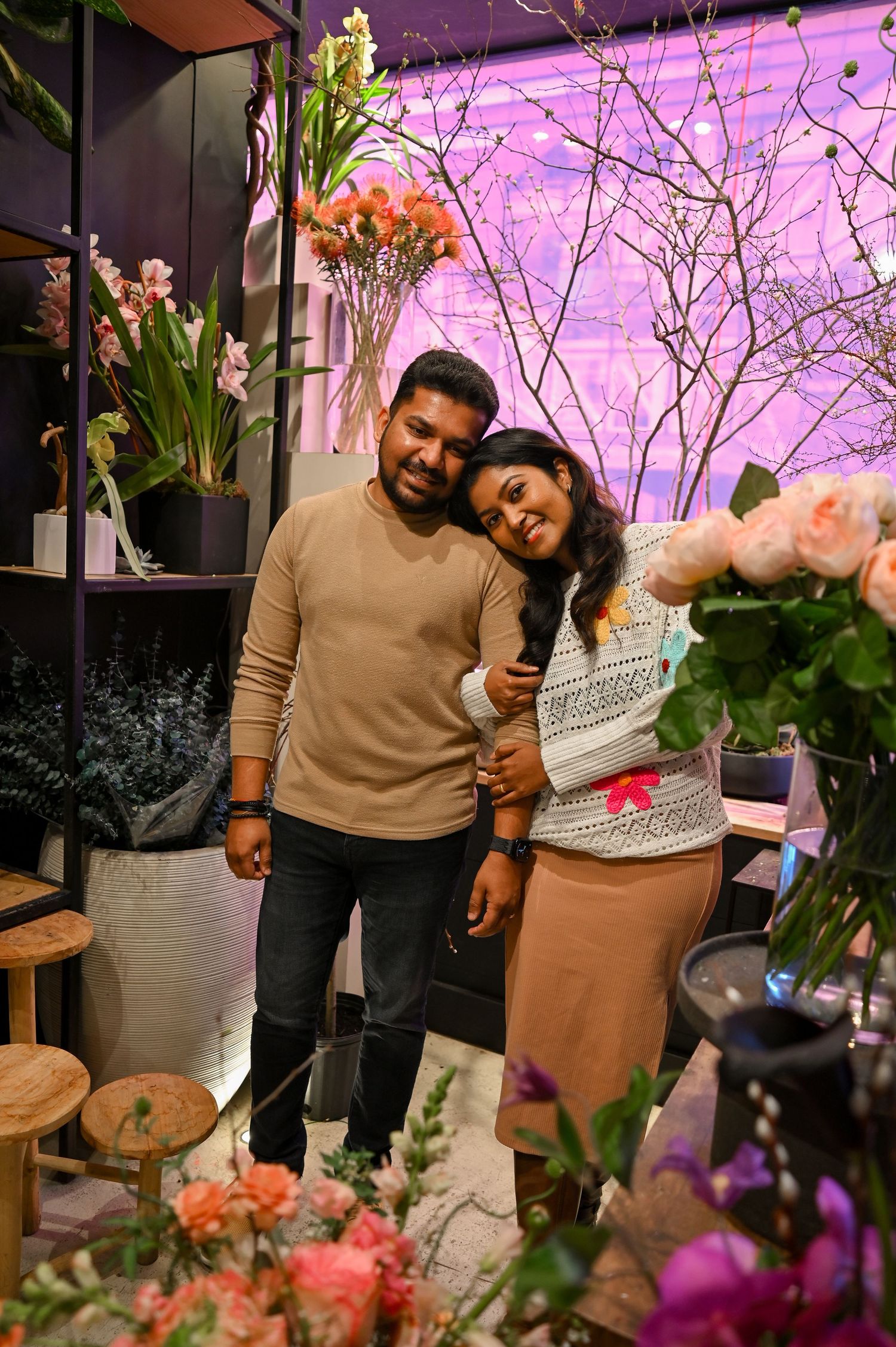 A couple poses together in a flower shop with pink lighting and floral displays.