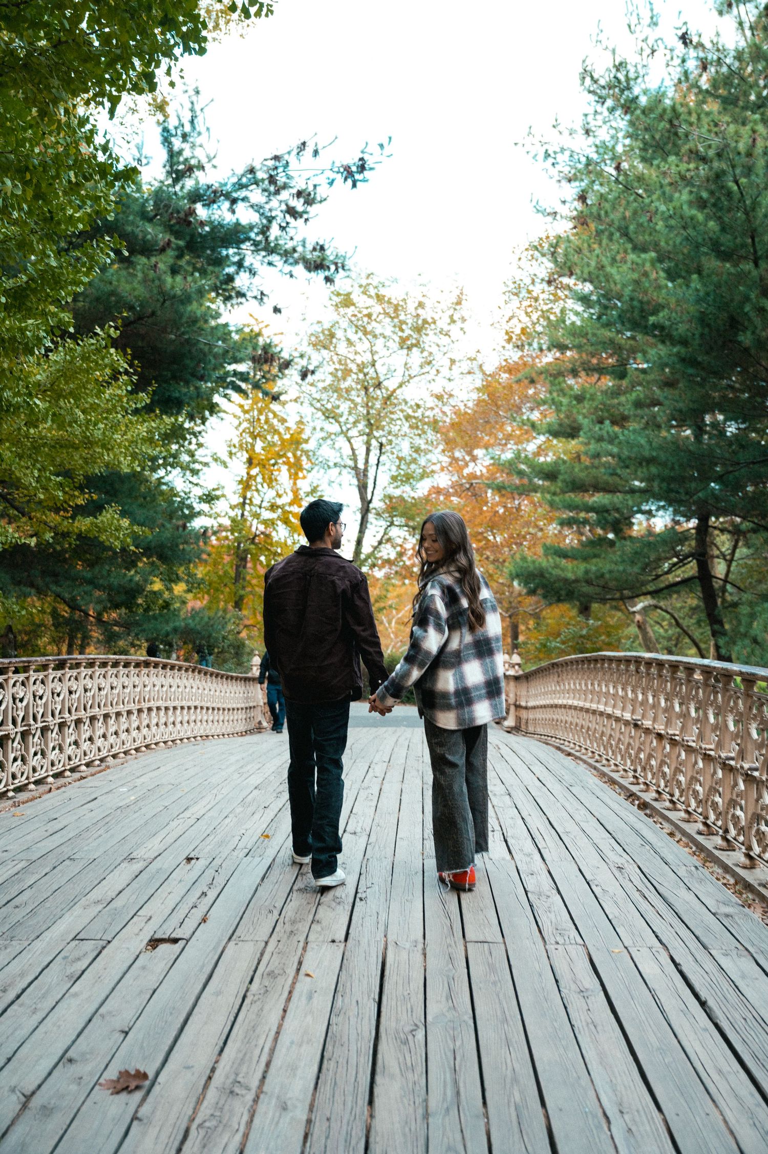 Two people walking hand in hand across a wooden bridge surrounded by autumn trees.