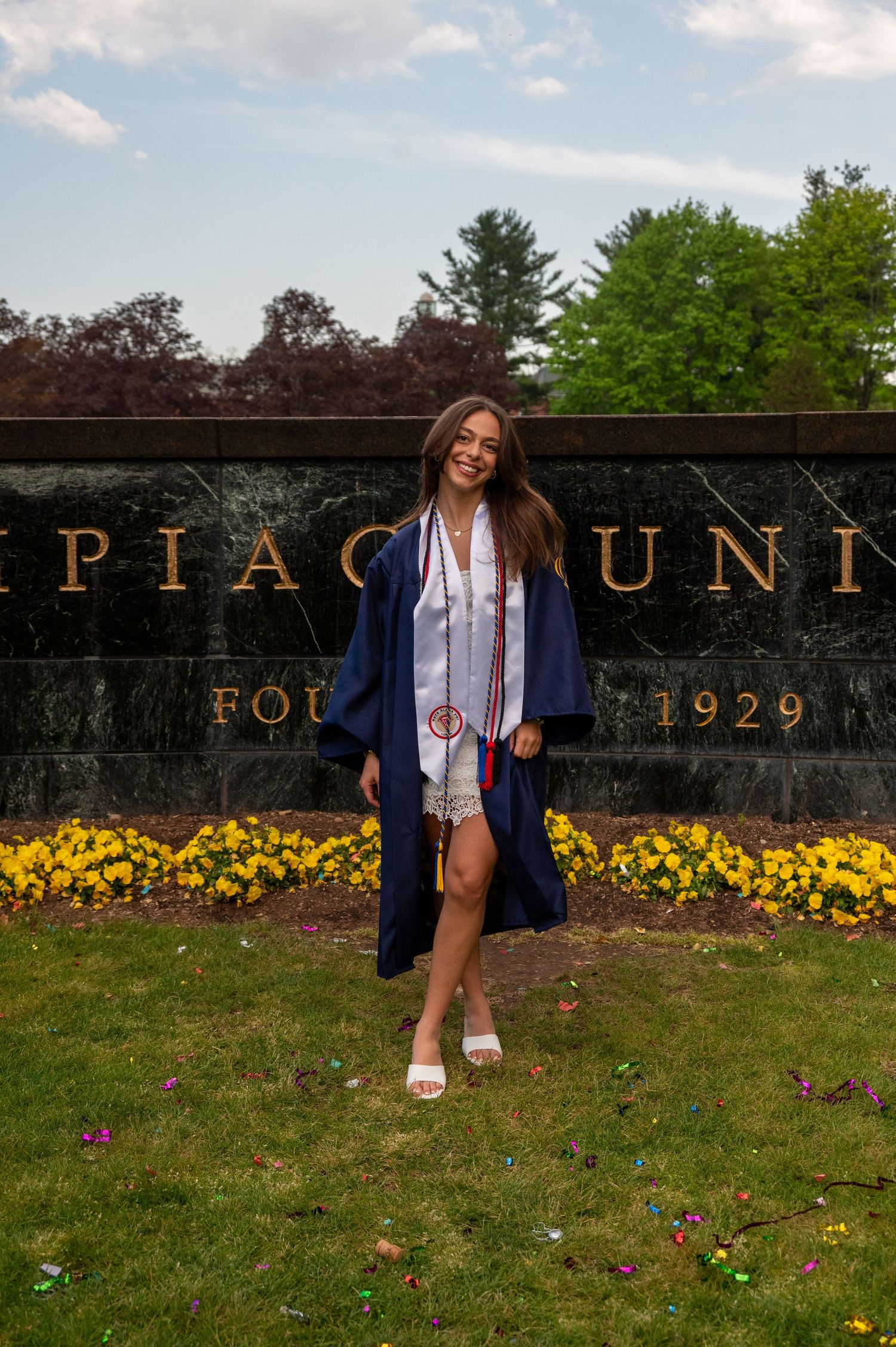 Graduate celebrates on campus lawn with yellow flowers and university signage.