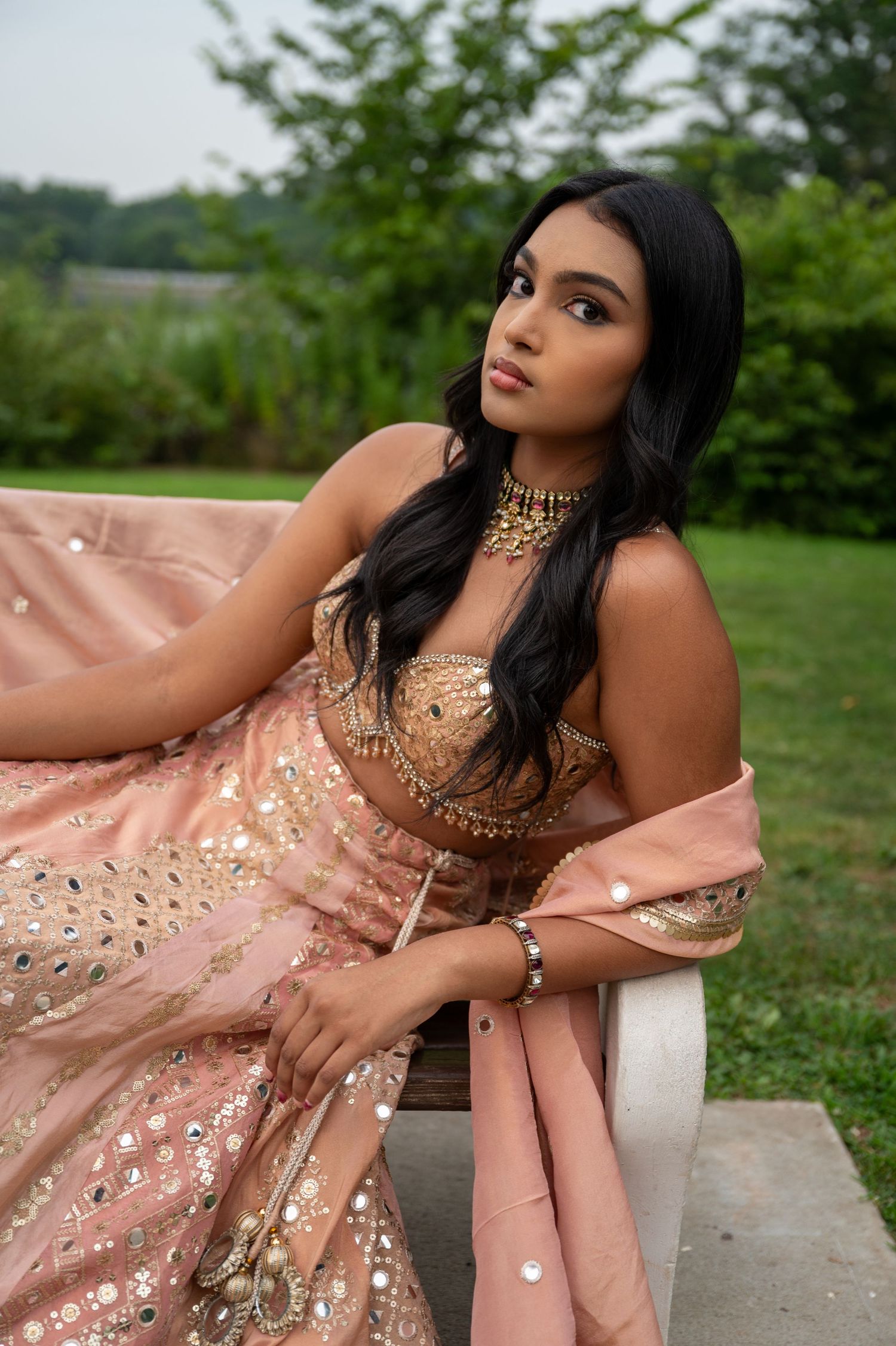 Model in peach embellished dress with gold jewelry poses gracefully outdoors.