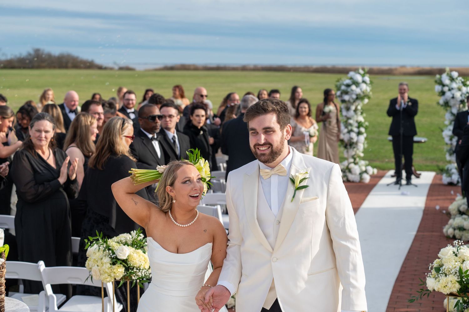 A wedding party celebrates outdoors on a sunny day with white floral arrangements and elegant formal attire.