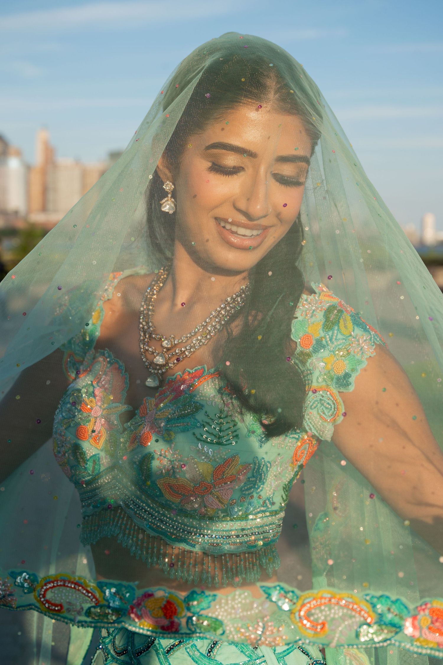 Woman in teal embroidered dress poses under sheer veil with city backdrop.