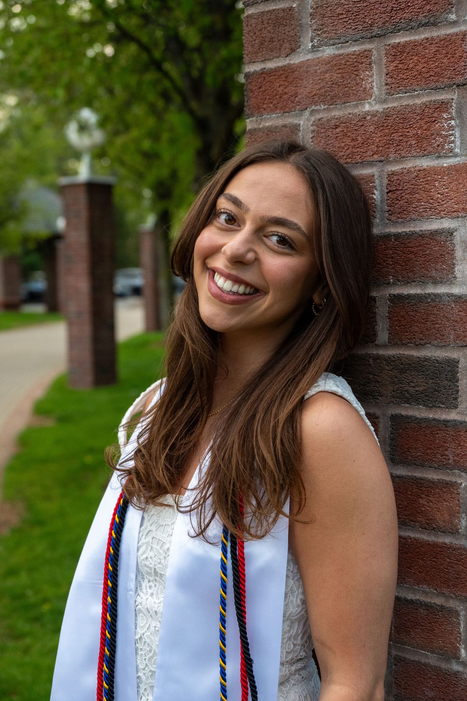 Graduate in white dress poses against brick wall with bright smile.