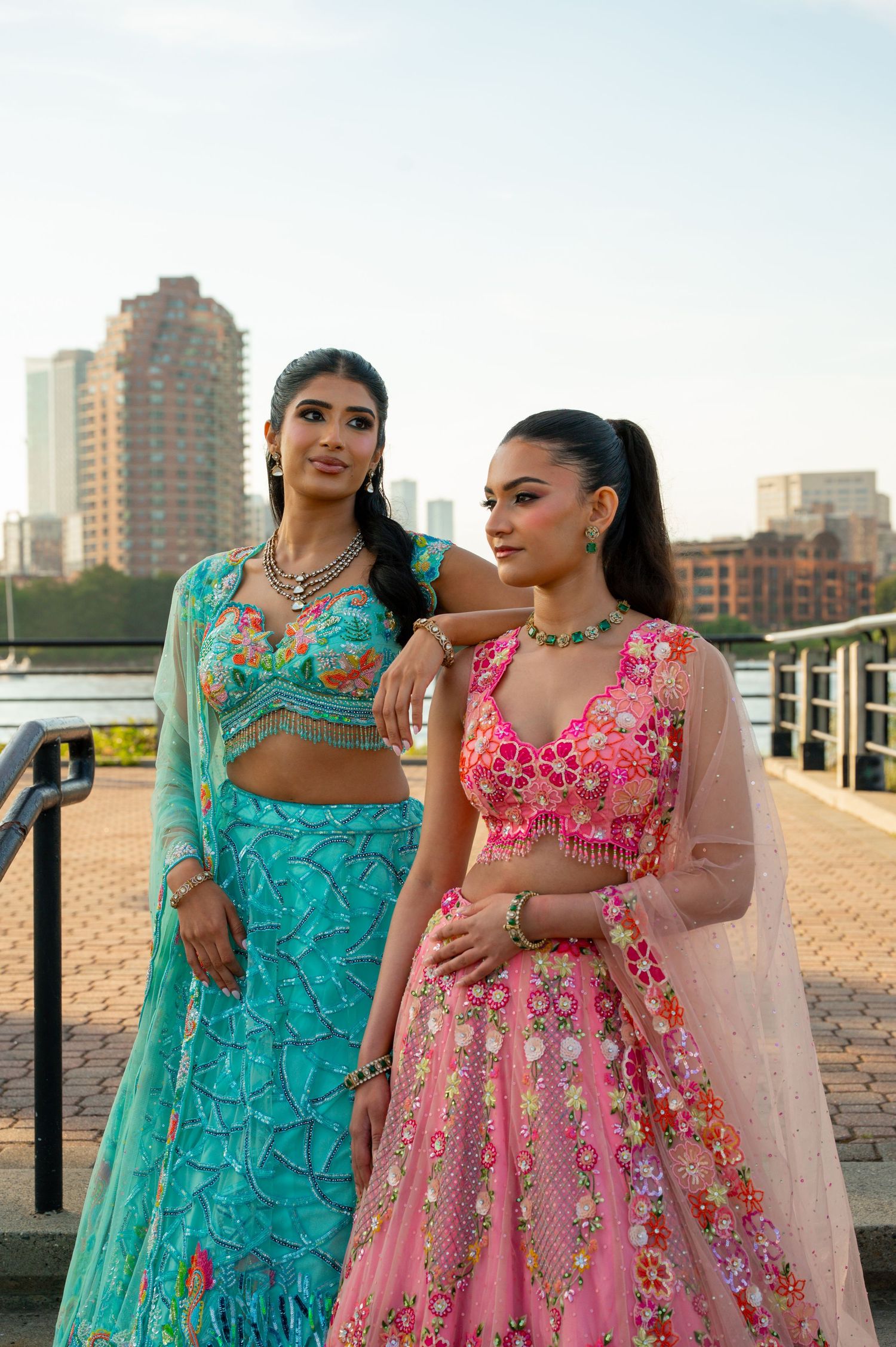 Two women in traditional Indian outfits pose together on brick pathway by waterfront.