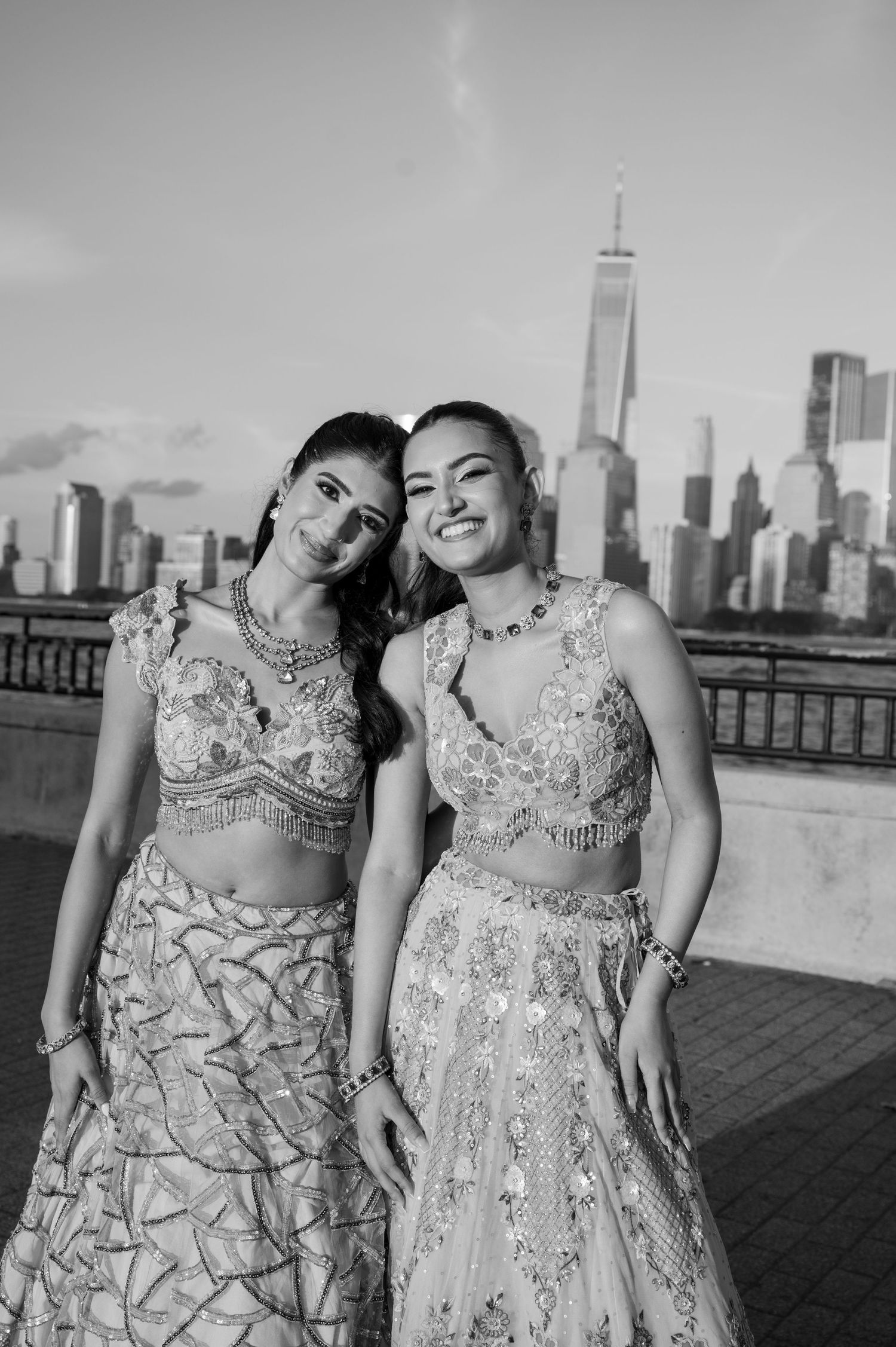 Black and white photo of two friends in embellished gowns sharing joyful moments by the waterfront.
