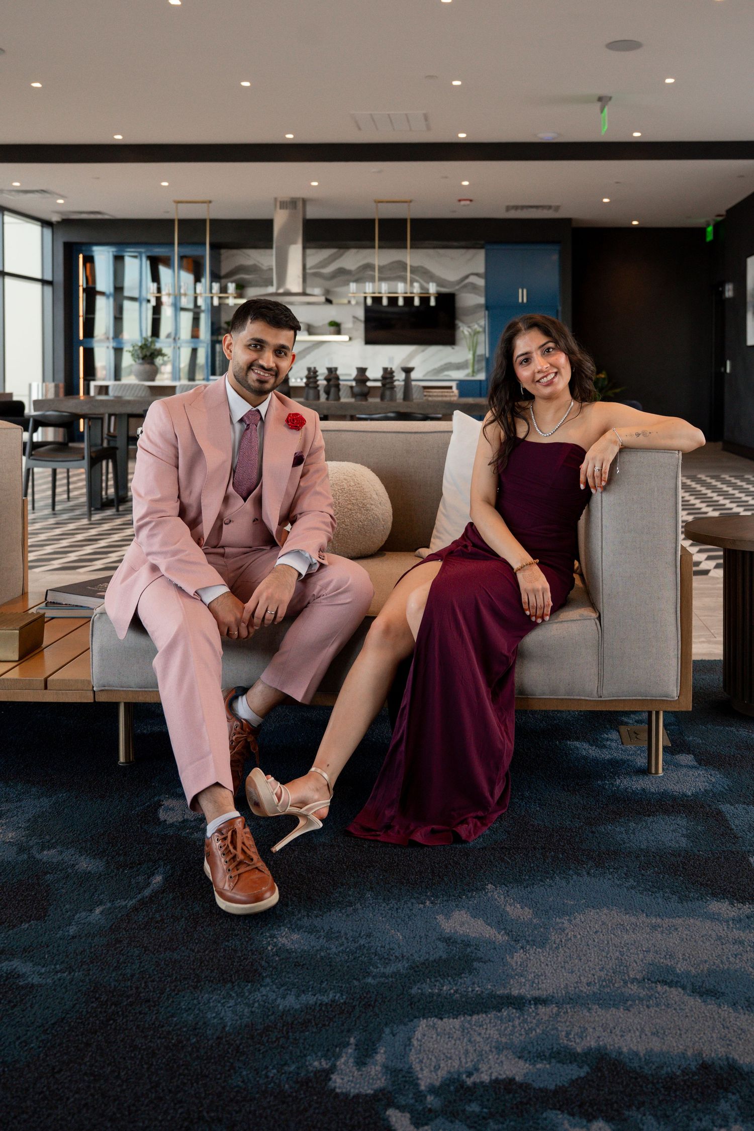 A couple in formal attire, with a pink suit and burgundy dress, pose playfully on a carpeted floor in an elegant venue.