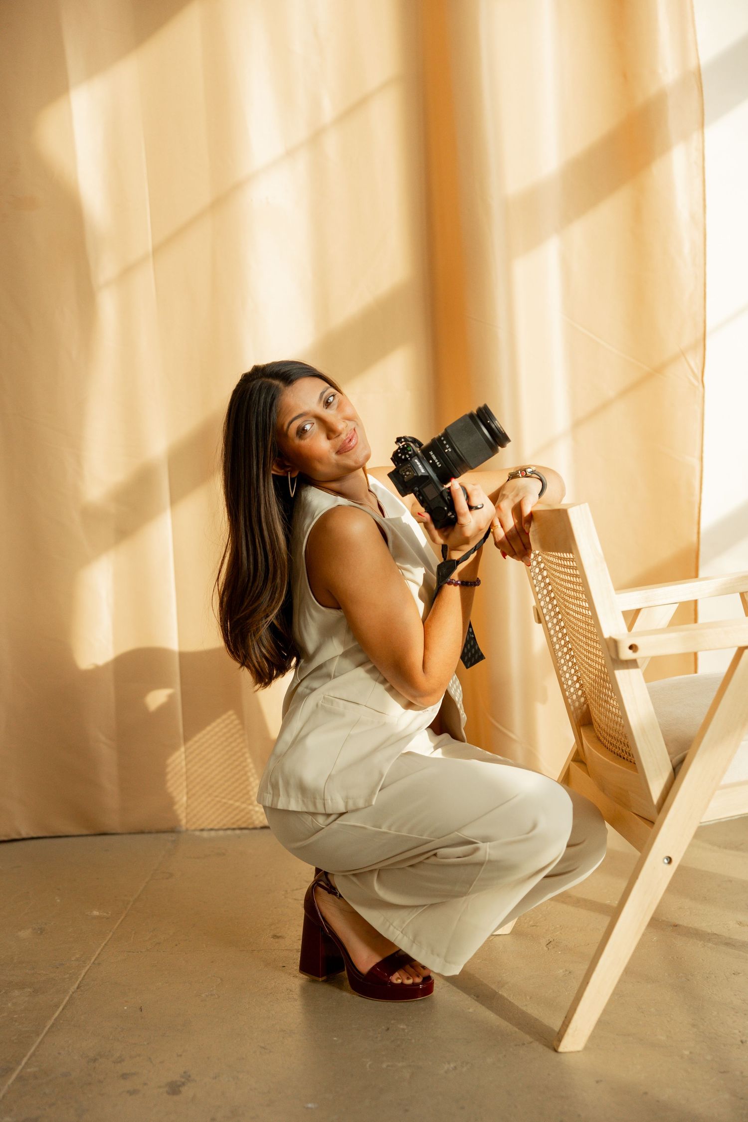 A photographer in a white dress crouches low while taking creative shots in a sunlit room with dramatic shadows.