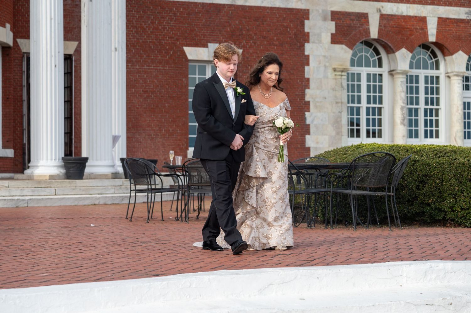 Wedding party members walk along a brick courtyard outside an elegant colonial-style building with white columns.