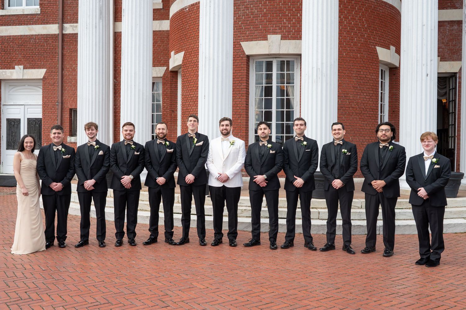 A line of formally dressed people in dark suits stand against a red brick building exterior.