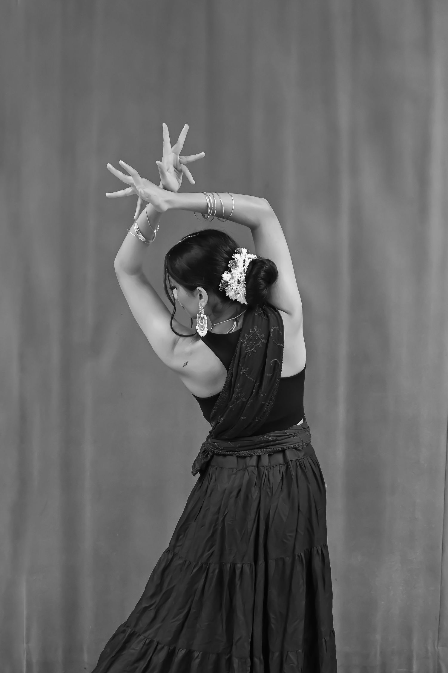A dancer in a black dress performs a graceful pose with raised arms against a gray backdrop in black and white.