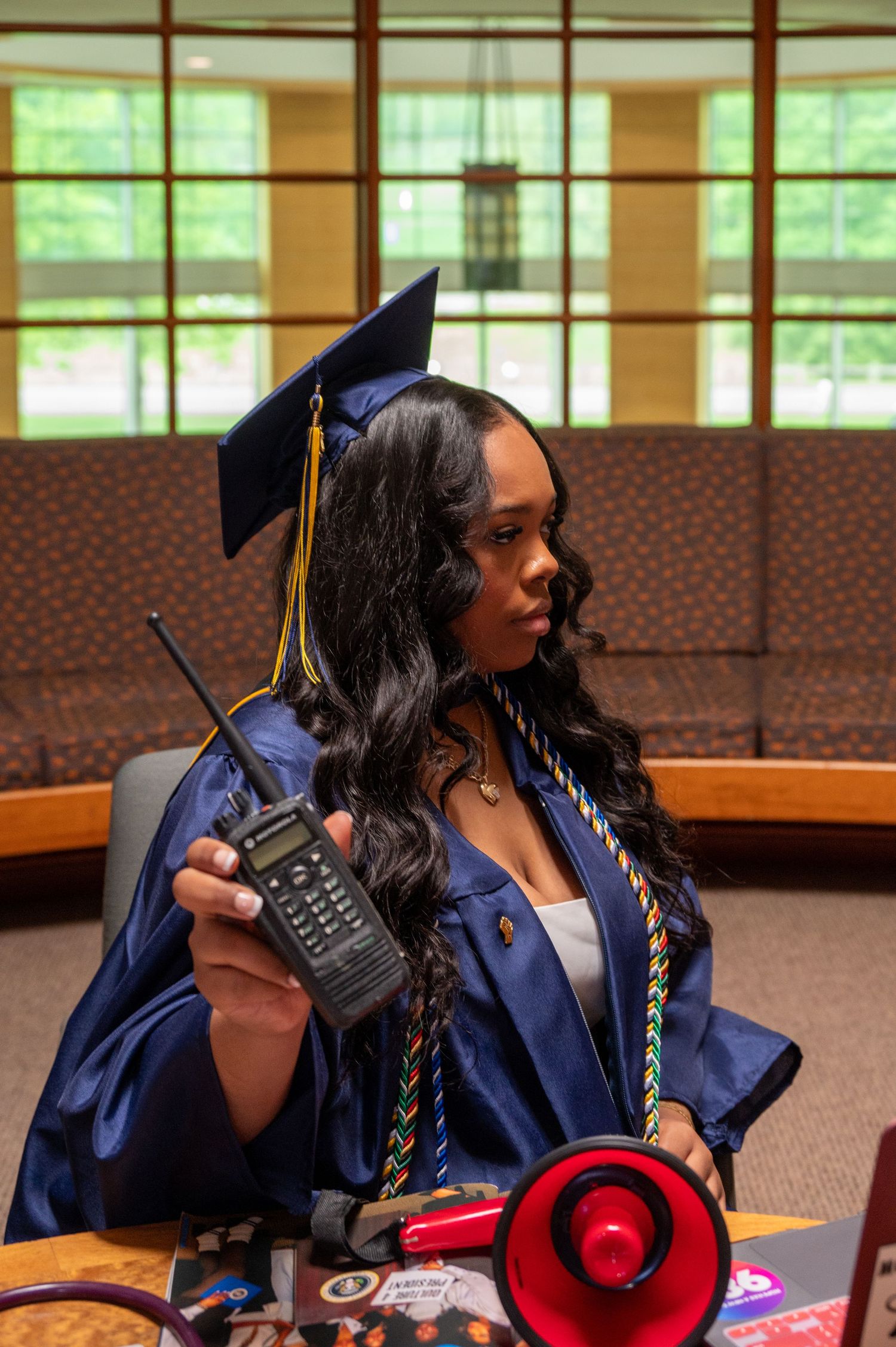 A graduate in navy blue regalia poses with academic accessories in a windowed room.