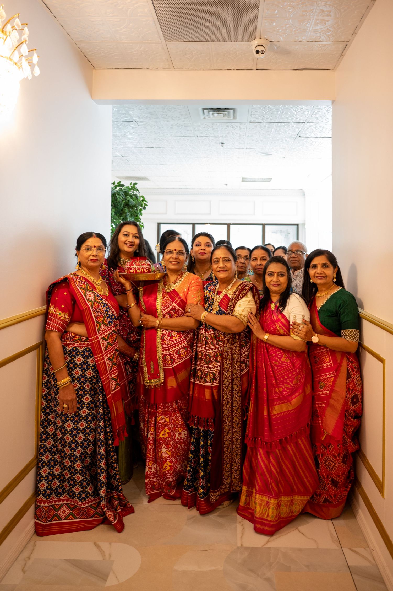 Group photo of women wearing traditional red and gold Indian wedding saris in a hallway.