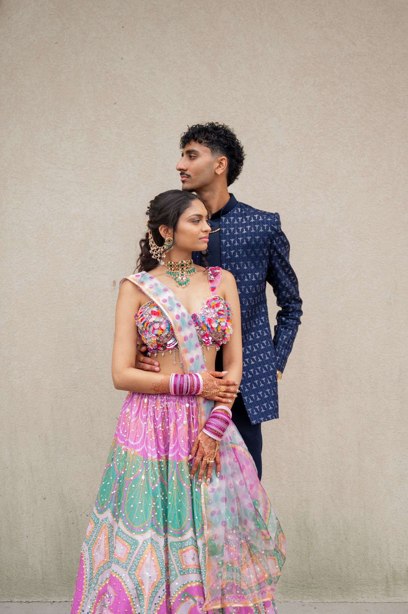 Couple in traditional Indian wedding attire, woman in colorful lehenga with jewelry, man in navy blue sherwani, posing together.