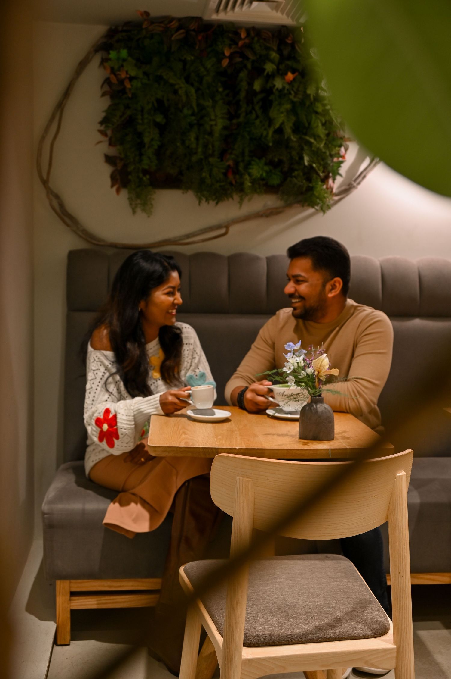 A couple enjoys coffee and conversation at a cozy cafe table under hanging plants.