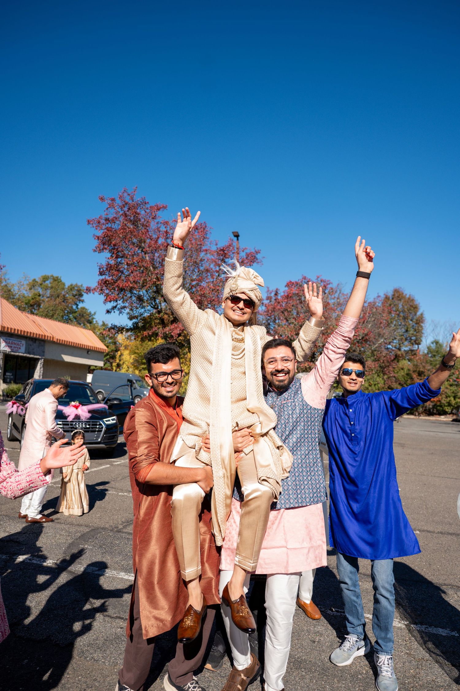 A group celebrates outdoors in traditional Indian attire with raised arms against a bright blue sky and autumn trees.