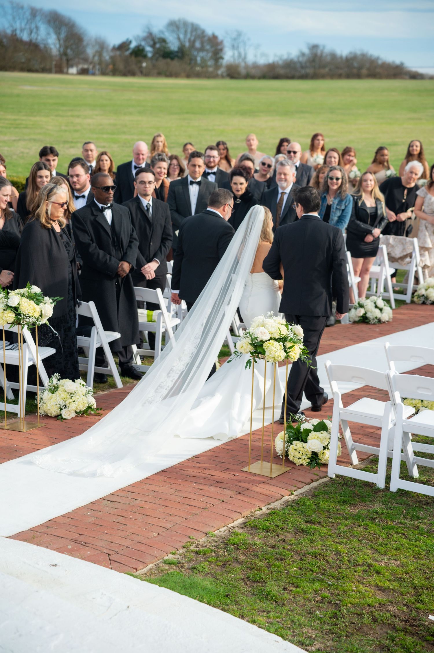 An outdoor wedding ceremony setup with white chairs and floral arrangements on a brick pathway.