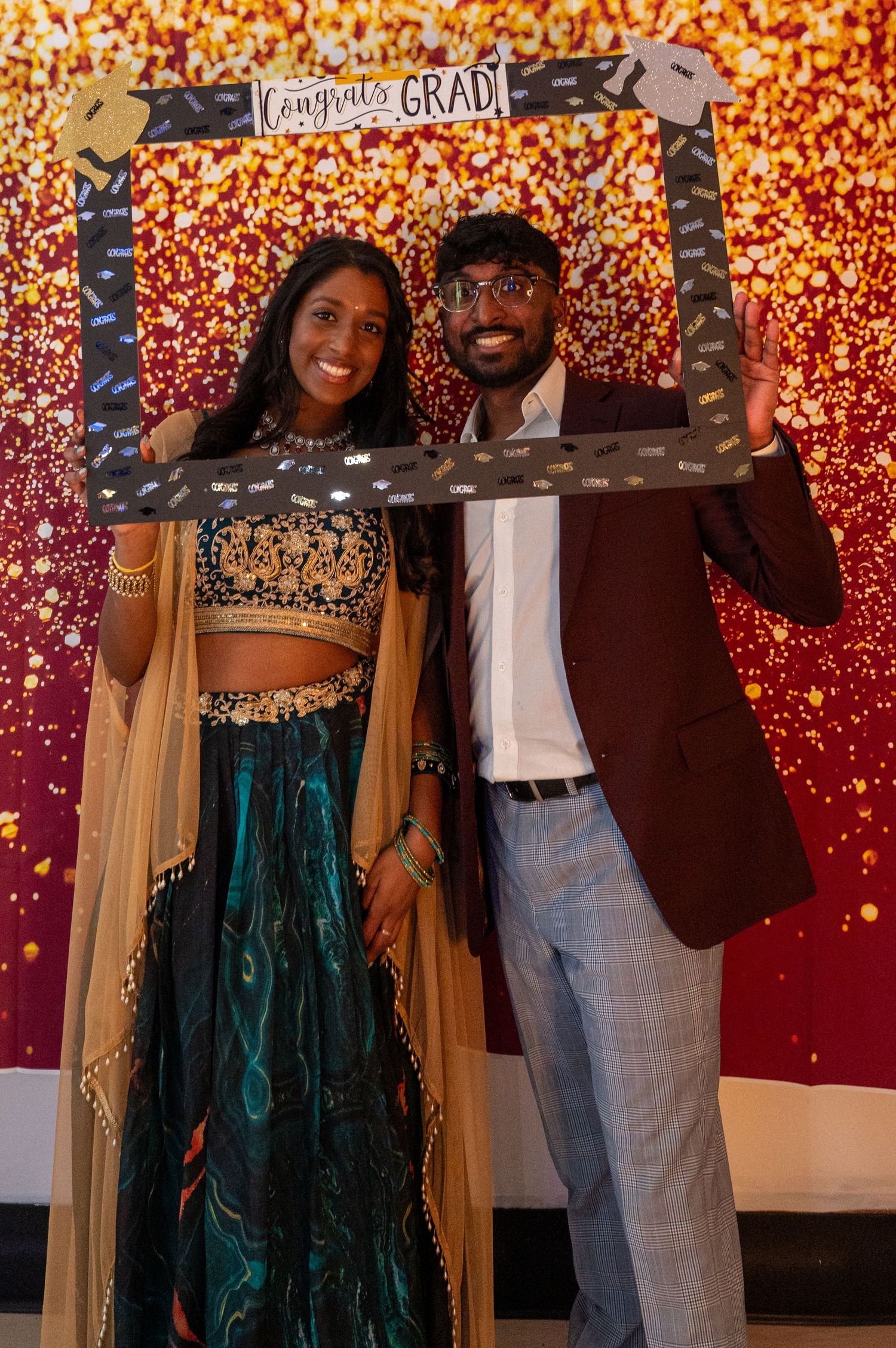 Couple posing with graduation-themed photo frame against sparkly backdrop.