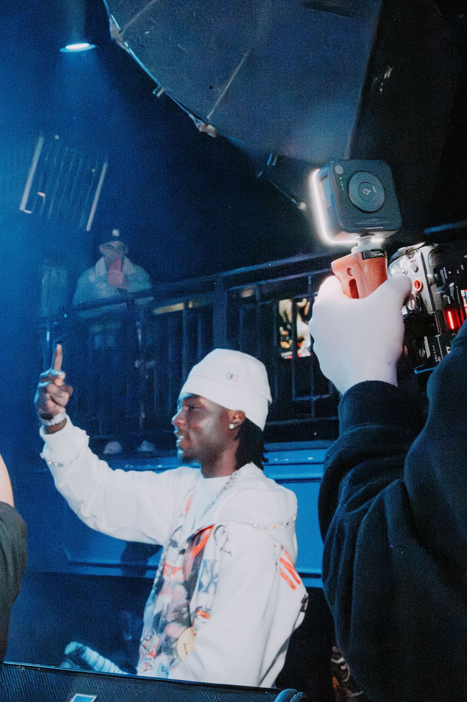 Person in white hoodie and baseball cap performing at a nightclub under blue lighting.
