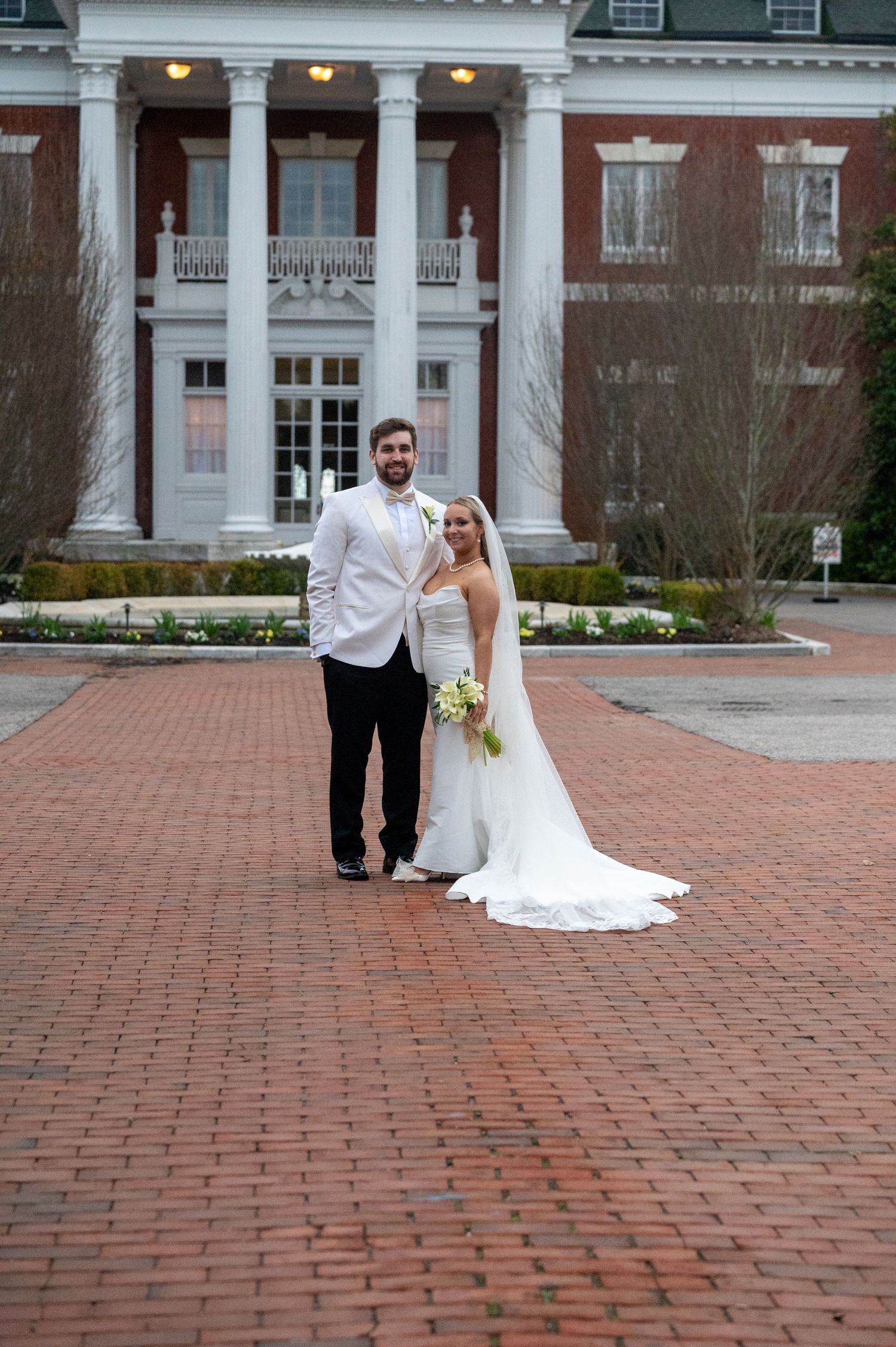 A wedding couple poses on a brick courtyard in front of a stately colonial building with white columns.