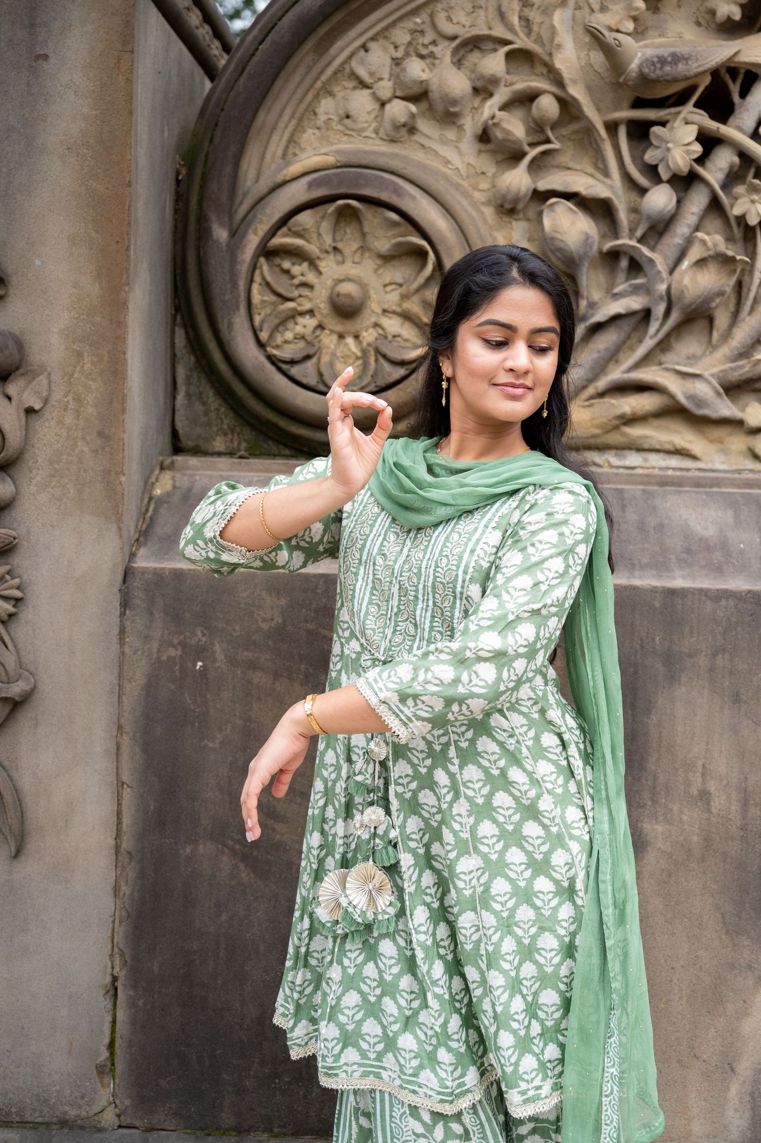 Person wearing elegant mint green embroidered outfit poses near ornate stone architecture.