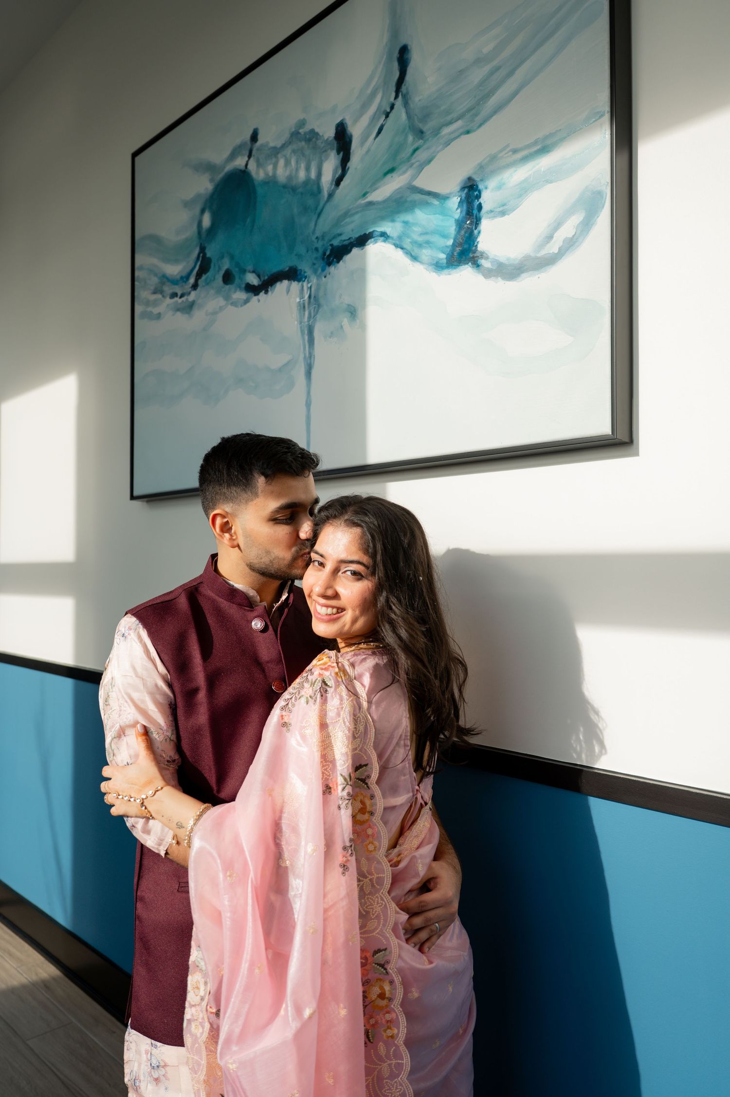 A romantic couple embraces near a wall with blue artwork, the person wearing a pink lace dress and other in burgundy attire.