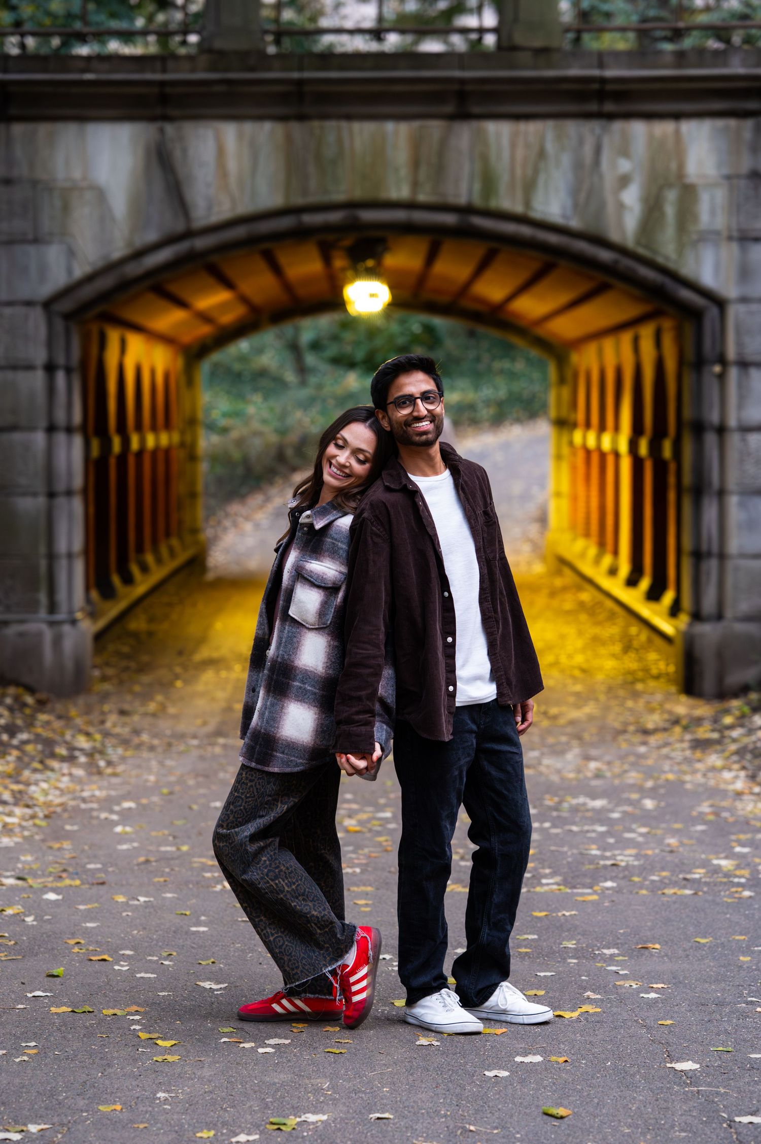 Couple in casual outfits posing playfully in front of an illuminated stone tunnel with autumn leaves.