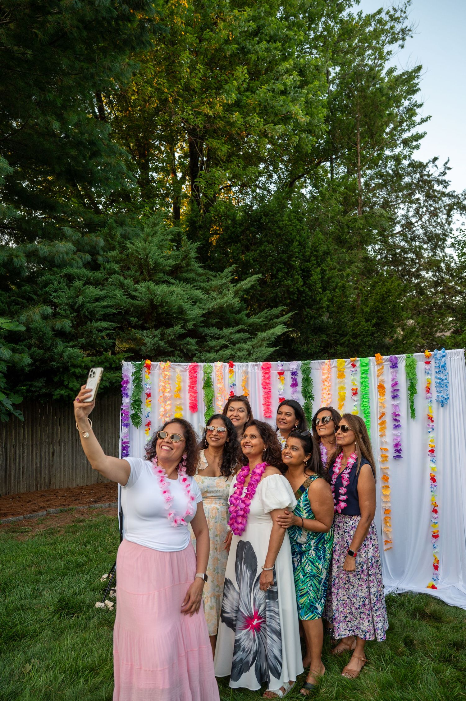 Friends posing for selfies at an outdoor party with colorful streamer decorations and floral backdrop.