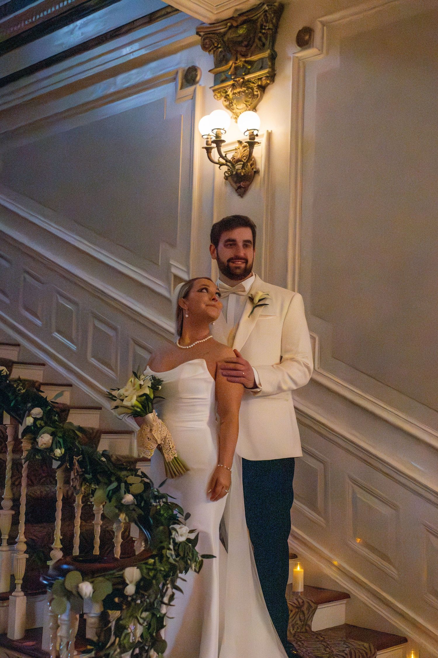 A couple poses together on an elegant staircase with white walls and ornate decor during what appears to be a wedding.