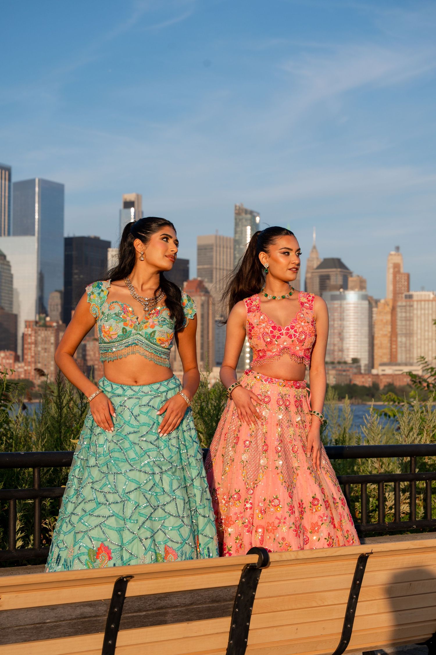 Models in complementary lehengas pose along waterfront railing with NYC skyline view.