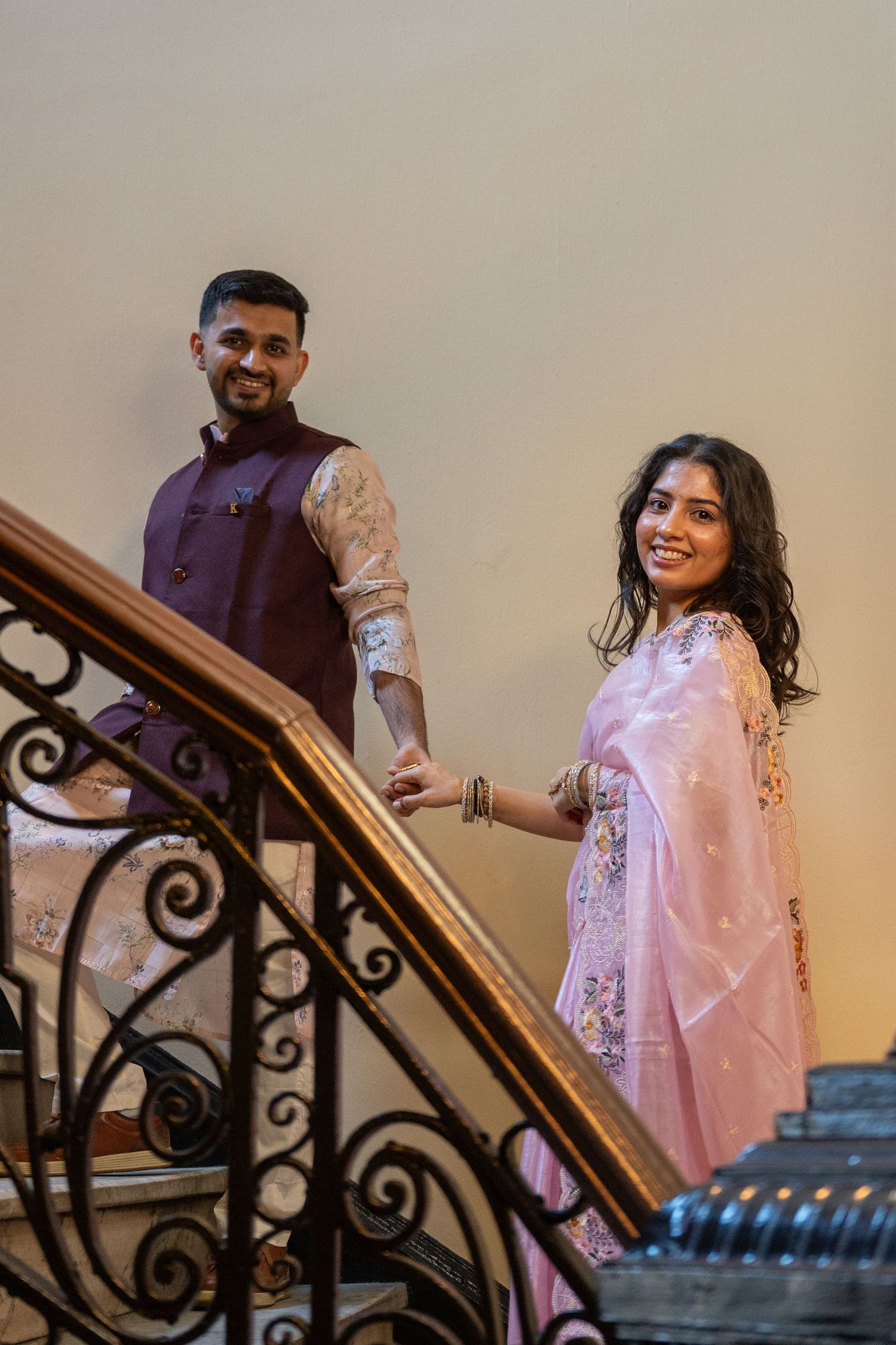 A couple in traditional Indian attire pose on an ornate staircase with wrought iron railings.