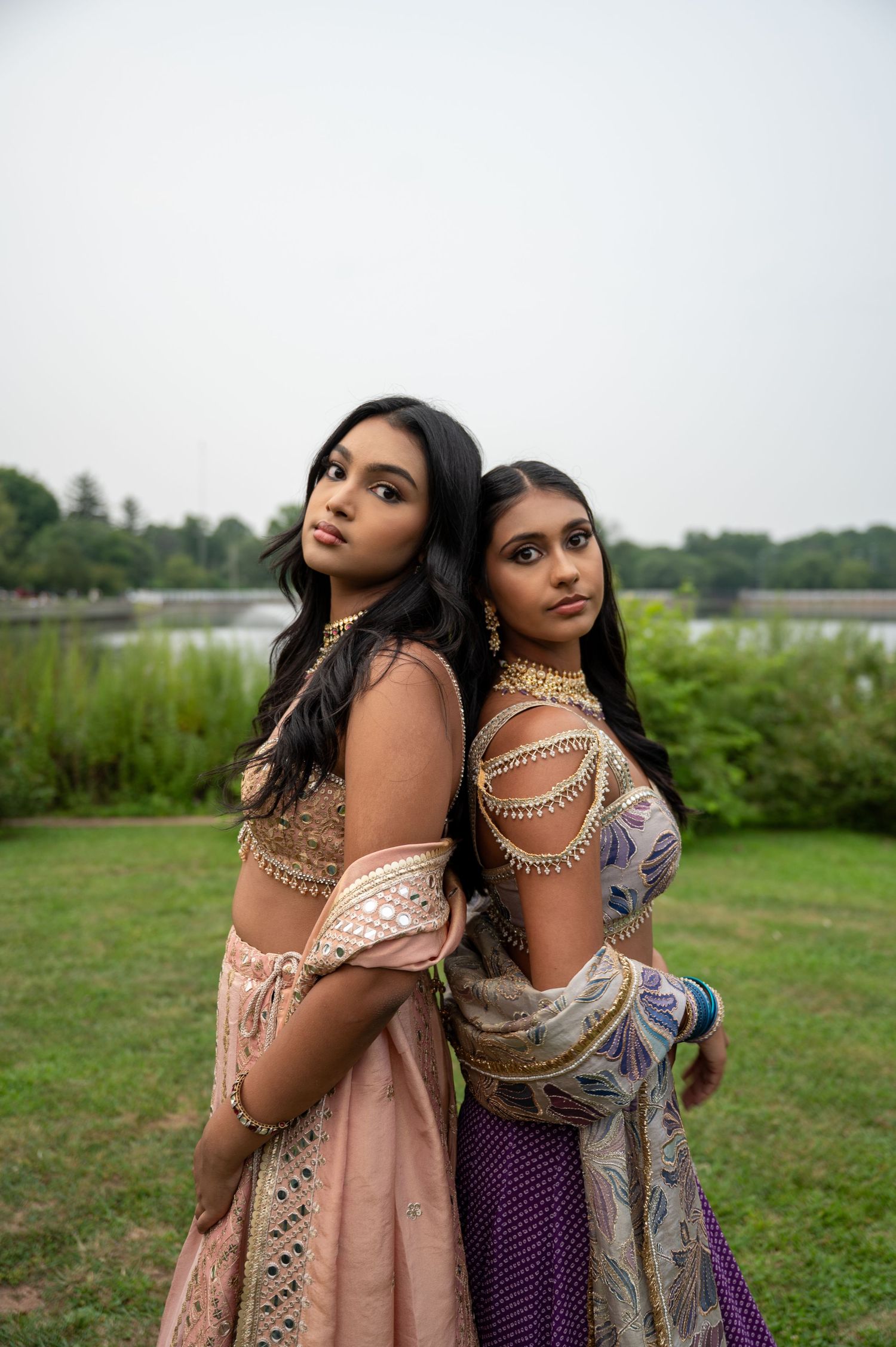Two women in traditional South Asian attire pose together in an outdoor setting with greenery in the background.