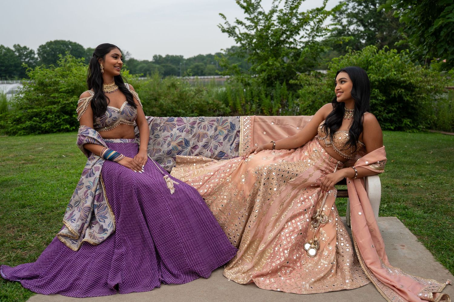 Outdoor fashion shoot featuring models in purple and peach formal gowns on grassy field.