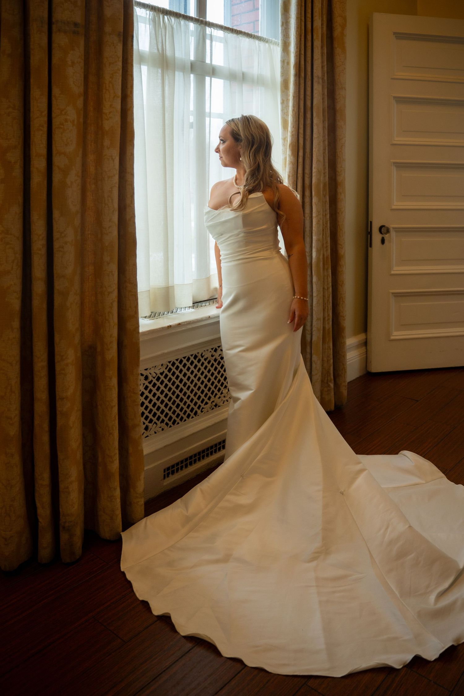 A bride in a flowing white wedding gown twirls elegantly by a sunlit window in a classic interior setting.