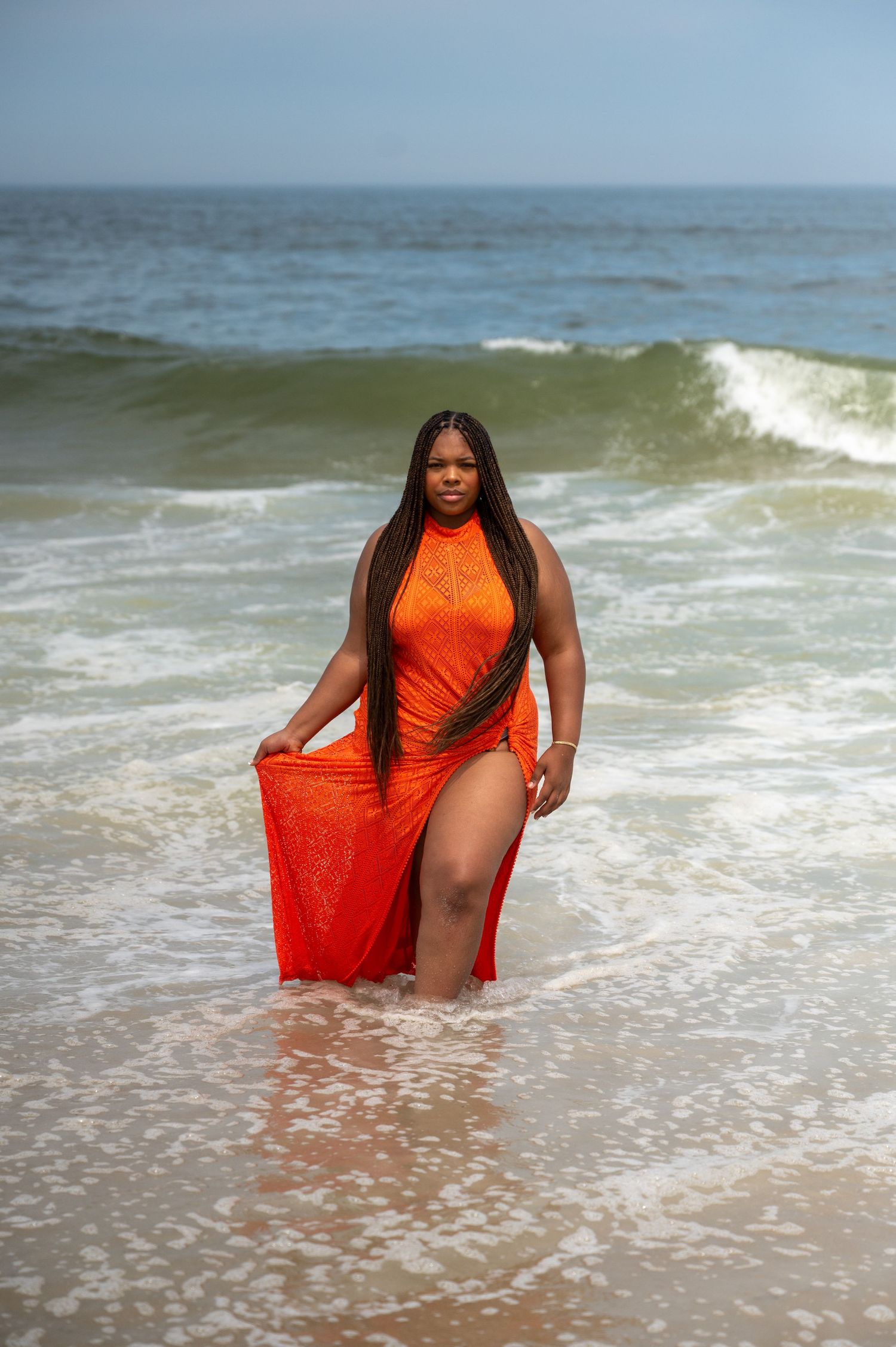 Figure in bright orange dress stands gracefully in ocean waves at beach.