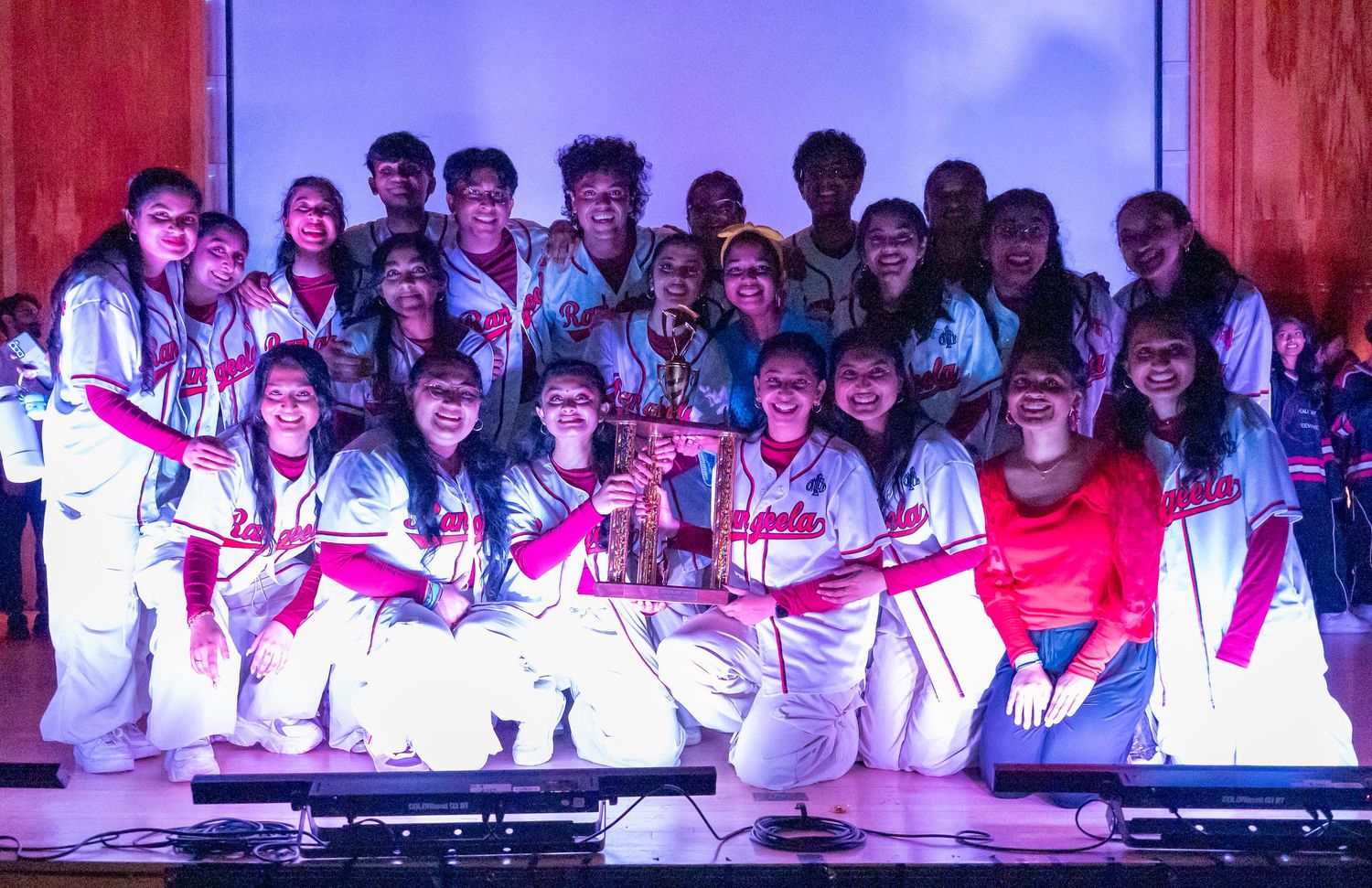 Performance group in white outfits poses together on stage under theatrical lighting.