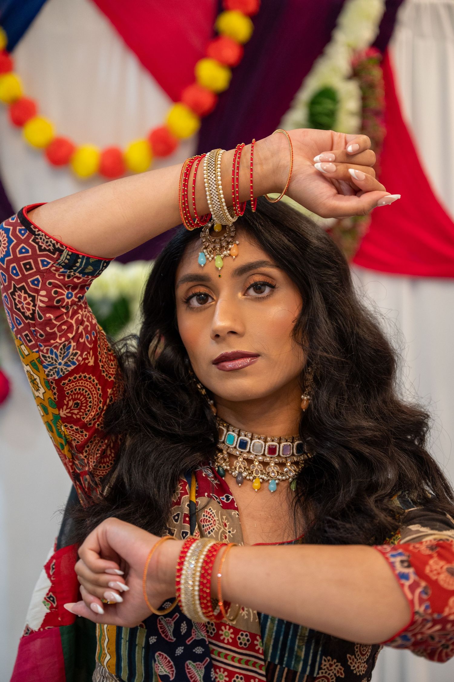 Woman in traditional Indian attire with jewelry and bindi posing against decorated backdrop.