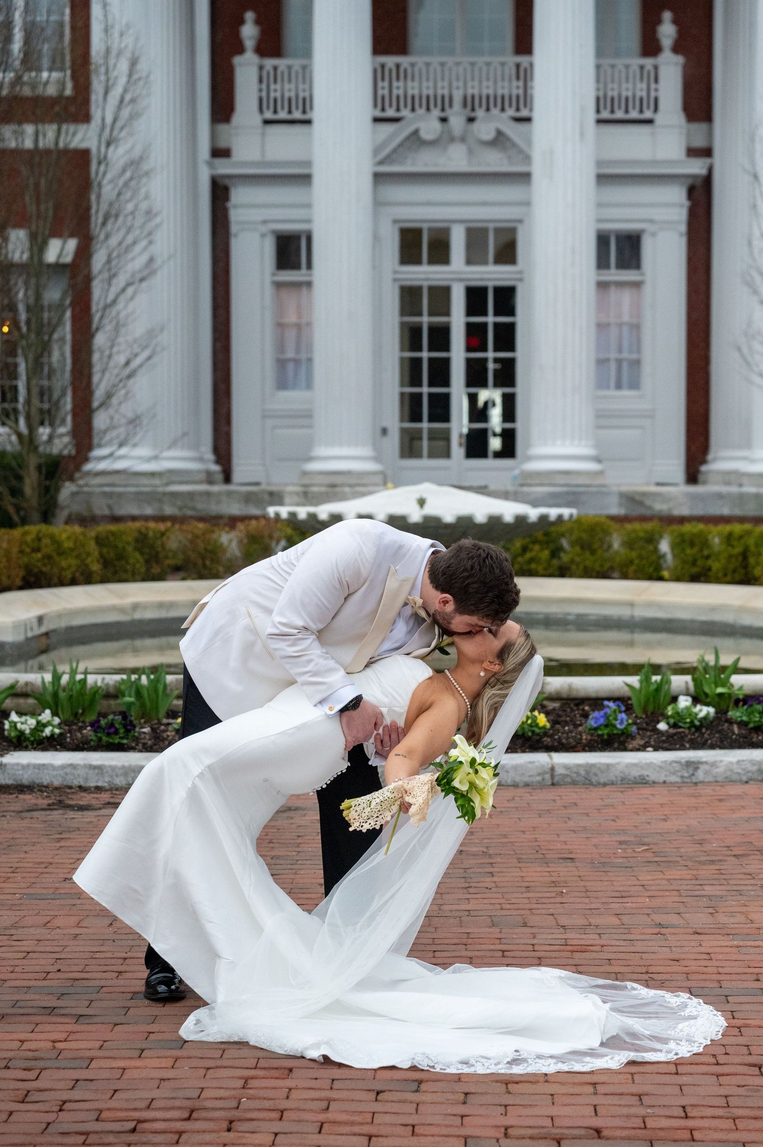 Wedding couple performs dramatic dip pose on brick courtyard in front of columned building.