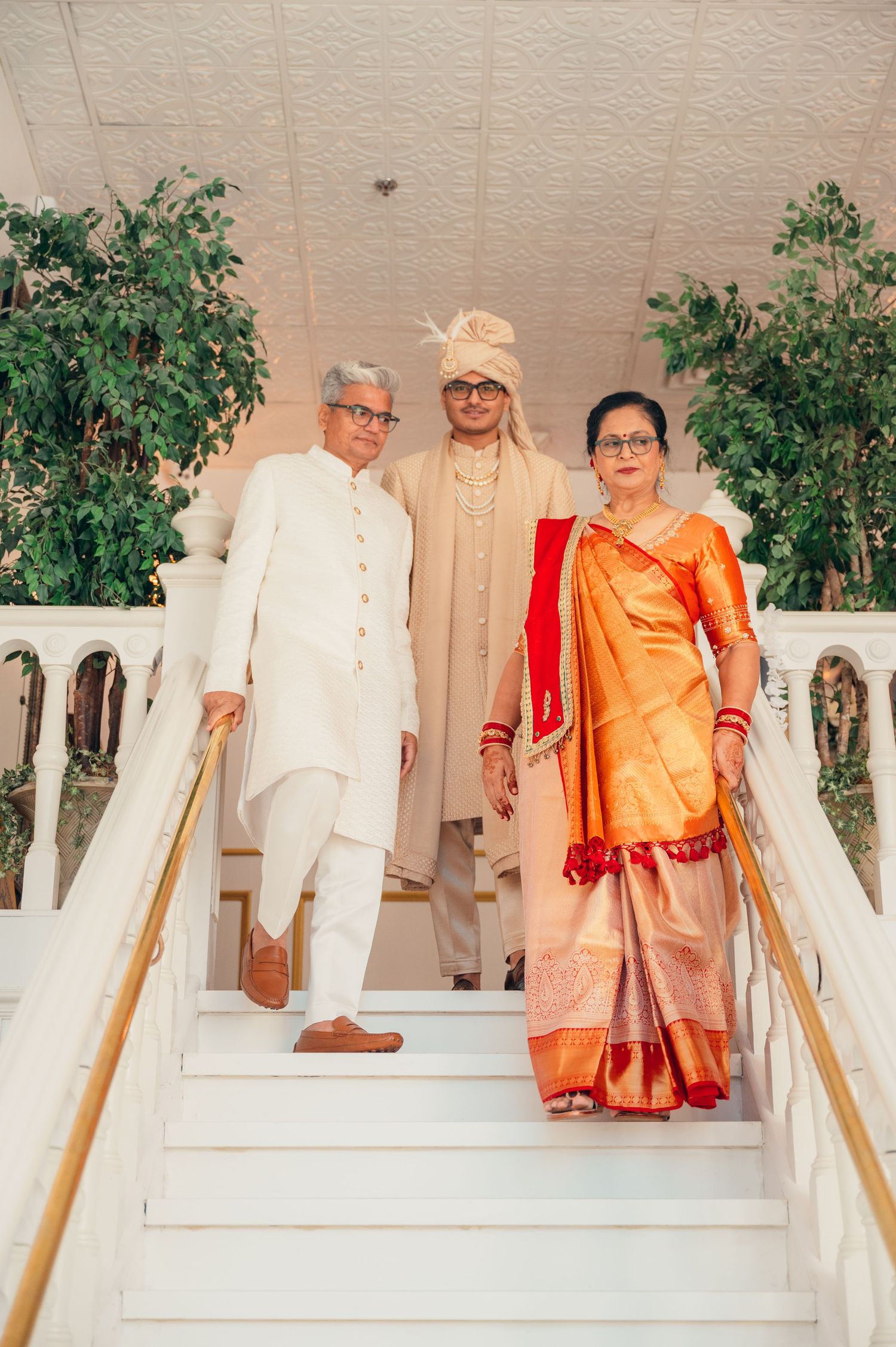 Groom and parents in traditional Indian attire descends white stairs during a wedding ceremony.