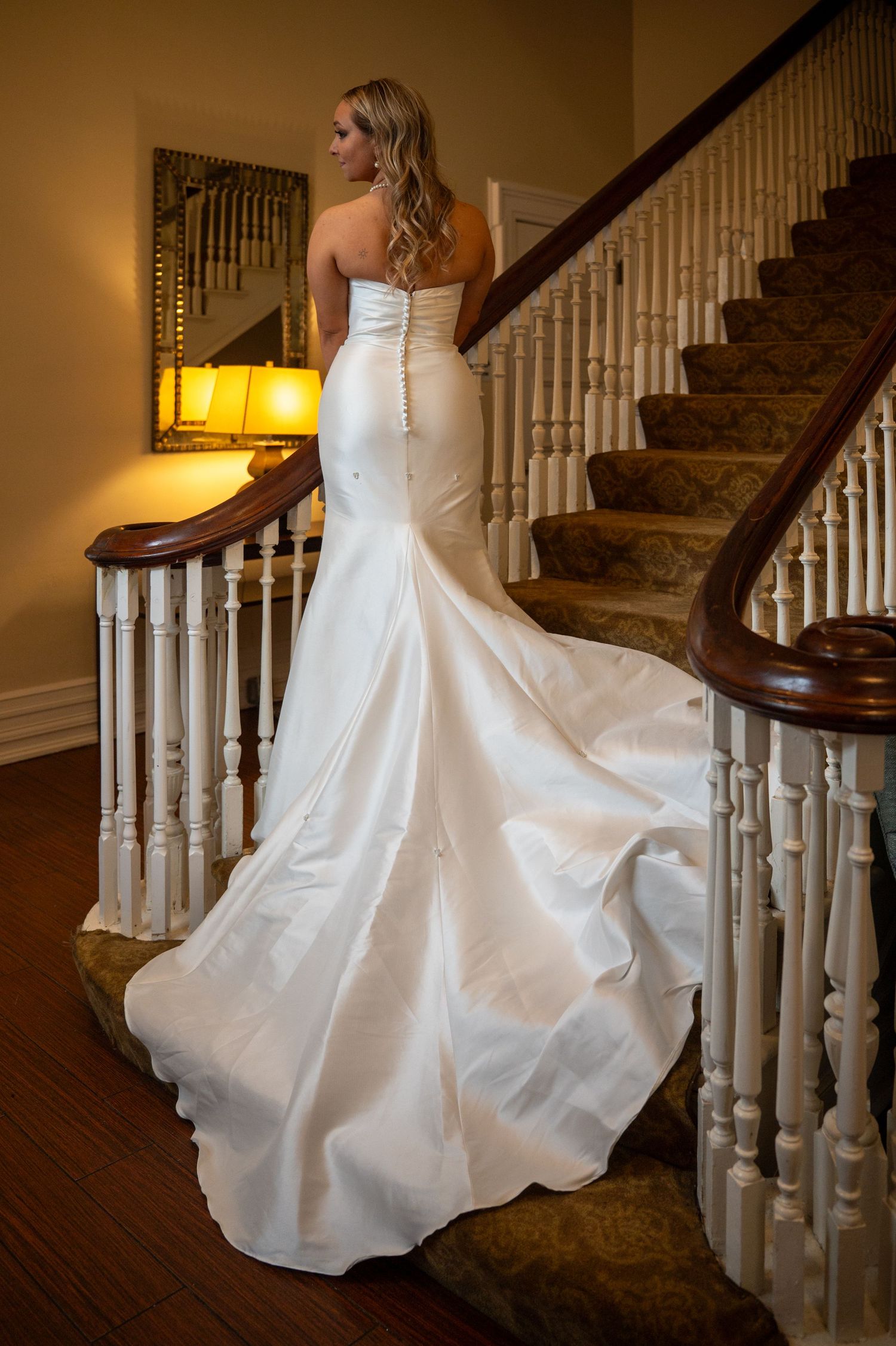 A white wedding dress twirls gracefully on a wooden staircase with white balusters and warm lighting.