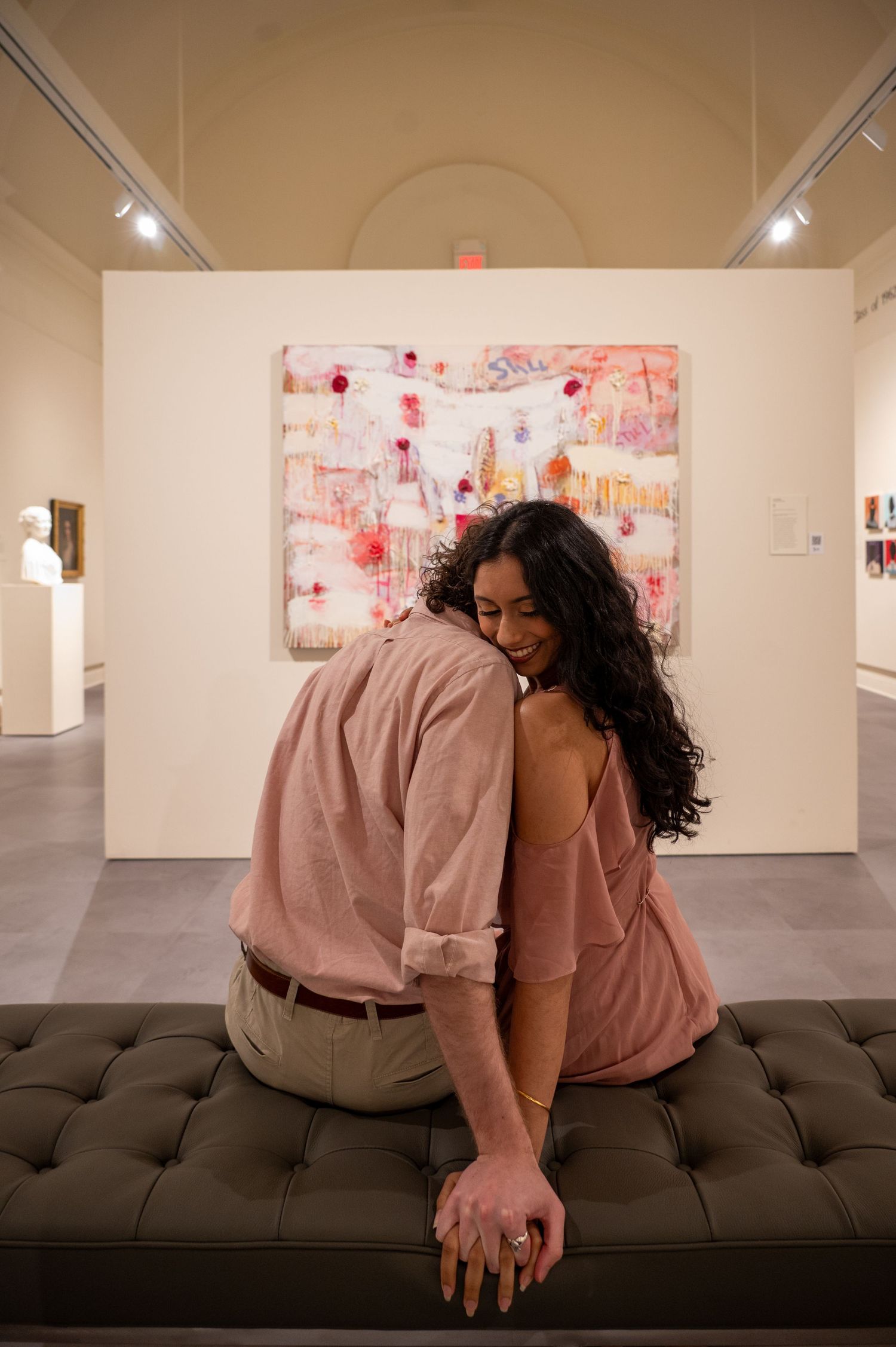 A couple sits together on a tufted bench in an art gallery with pink abstract artwork.